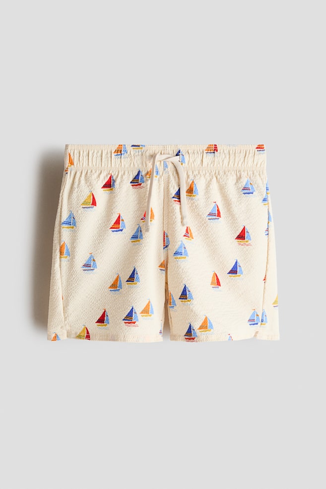 Vista frontal de shorts de baño color crema, con una tela texturizada y un patrón colorido de veleros. Están diseñados con una cintura elástica fruncida y un cordón blanco atado al frente, con pequeñas aberturas en el lado exterior de cada dobladillo. - 1