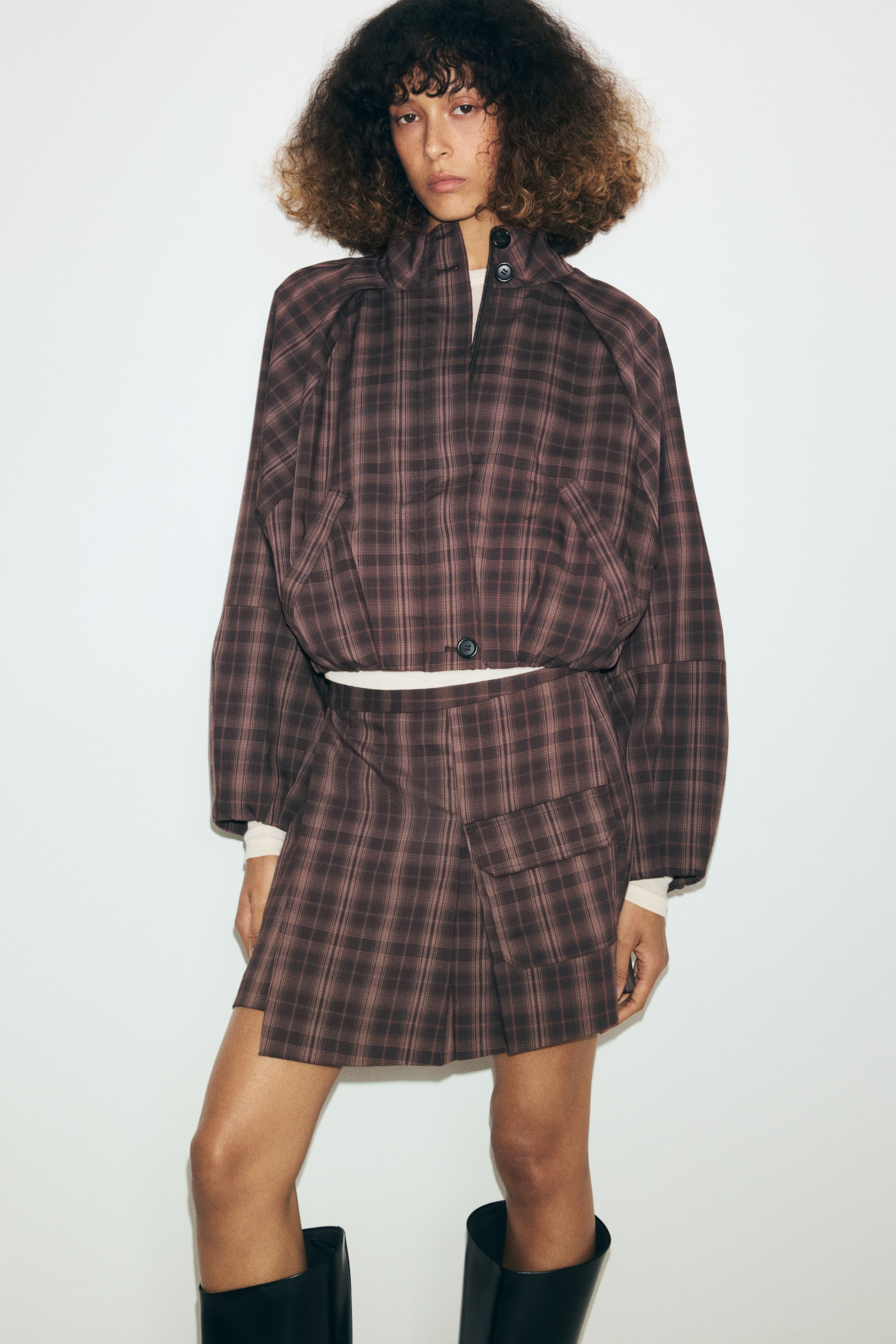 Ampliar la imagen: La modelo lleva puesta una chaqueta corta a cuadros marrón, con cuello mao, tapeta cortaviento con botones y mangas raglán. La chaqueta cropped se lleva sobre una prenda superior de color claro y se combina con una minifalda a cuadros del mismo estampado.