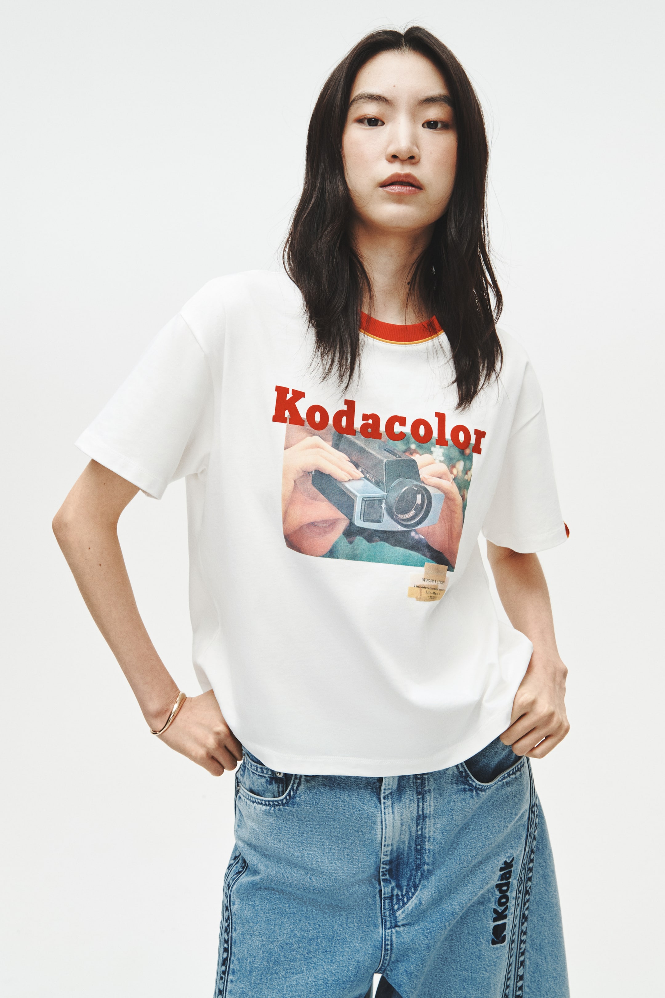 View larger image: A white t-shirt is showcased facing forward, featuring a red ribbed crew neck with a slim yellow stripe and matching red ribbed short sleeve cuffs. The front of the t-shirt displays a large graphic print with "Kodacolor" in red lettering positioned above an image of hands holding a vintage film camera, accompanied by a small rectangular graphic element below.