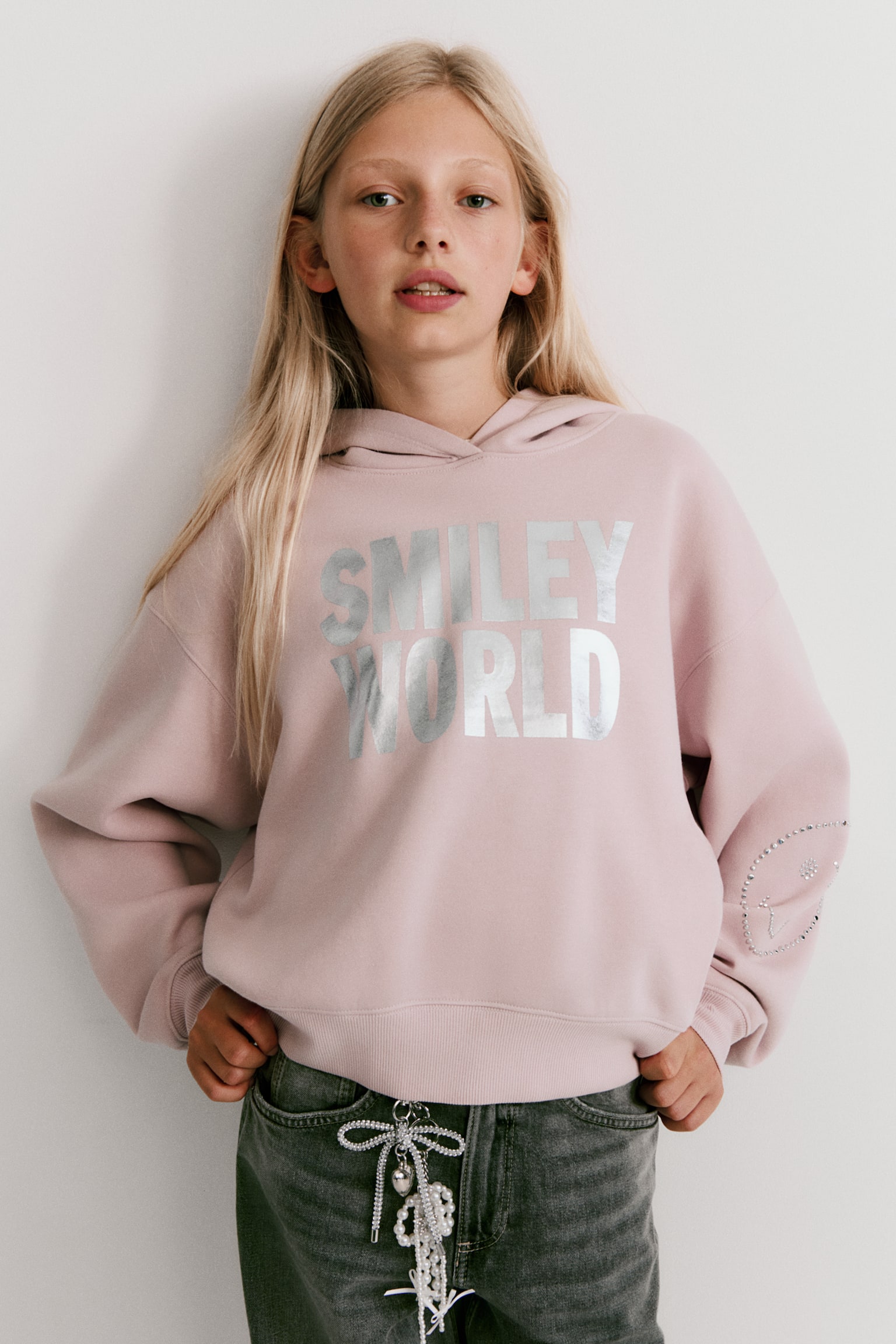 Um hoodie rosa apresenta uma gráfica metálica prateada "SMILEY WORLD" na parte frontal. Também exibe um design de cara sorridente com pontos prateados na manga esquerda e termina com punhos canelados e uma bainha canelada. - 1