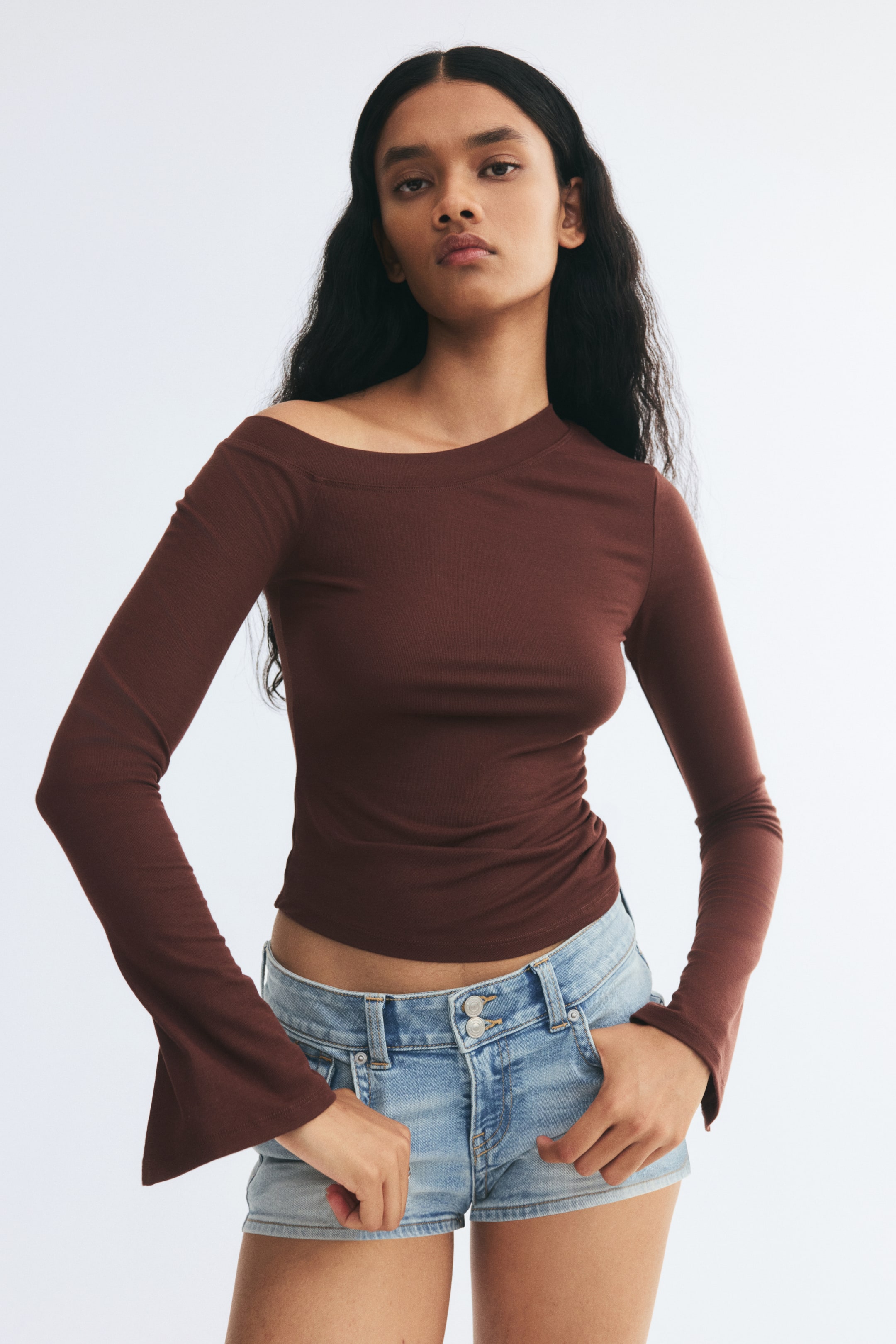 View larger image: The model is wearing a fitted dark brown long-sleeved top with an asymmetrical neckline that exposes one shoulder. The top features flared cuffs with slits and is styled with light-wash denim shorts.