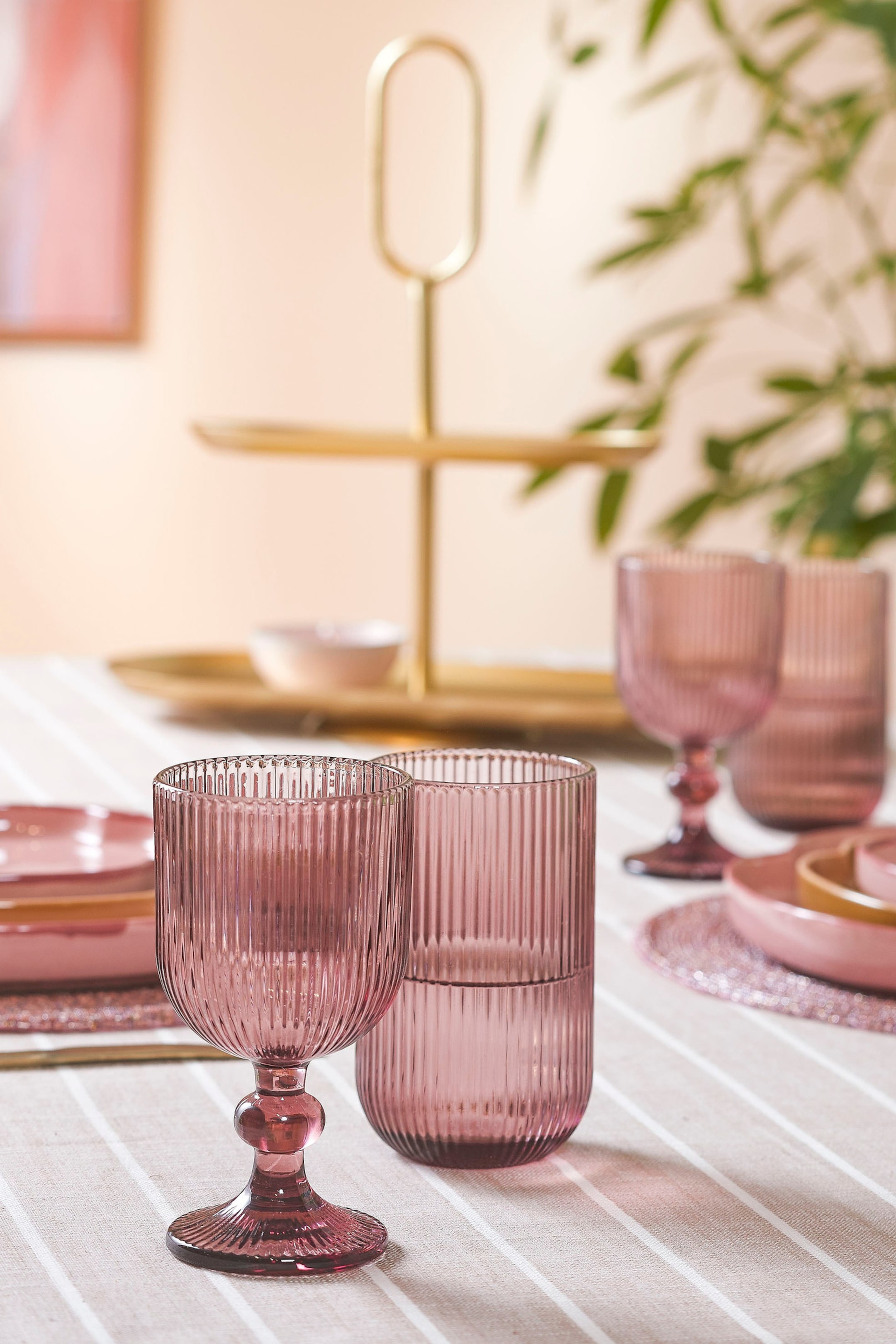 Grotere afbeelding bekijken: Een roze geribbelde kelk op een voet staat naast een bijpassende rechte geribbelde tumbler. De kelk heeft een bolvormig detail op de voet, terwijl de tumbler gevuld is met vloeistof.