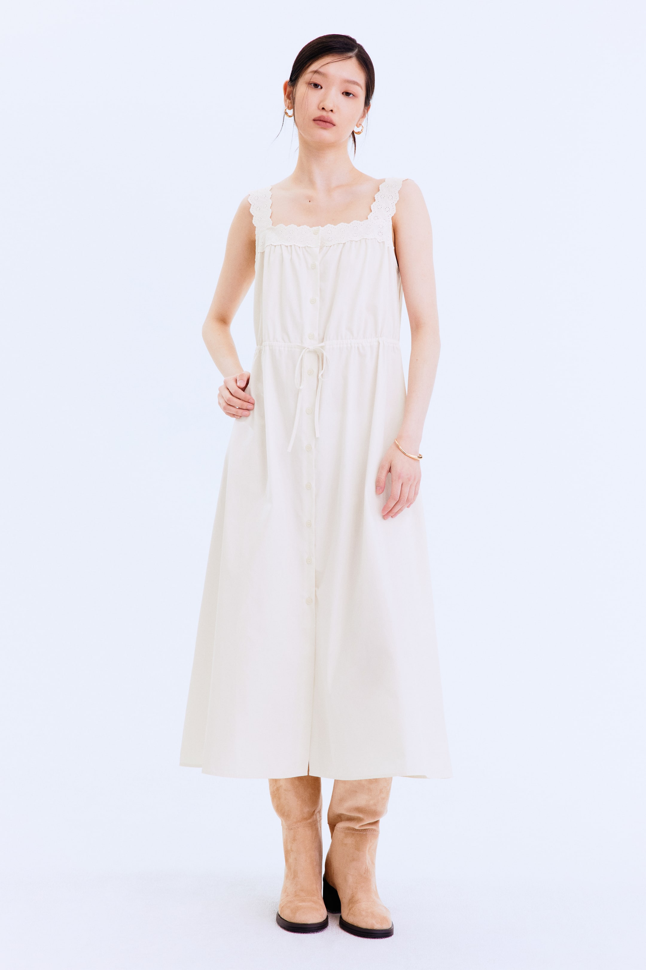 이미지 확대 보기: 오프 화이트 색상의 민소매 미디 드레스는 레이스 트림이 있는 스퀘어 넥라인과 앞면 전체에 버튼 플래킷이 특징입니다. 또한, 여유로운 A라인 실루엣을 잡아주는 드로스트링 허리끈이 돋보입니다.