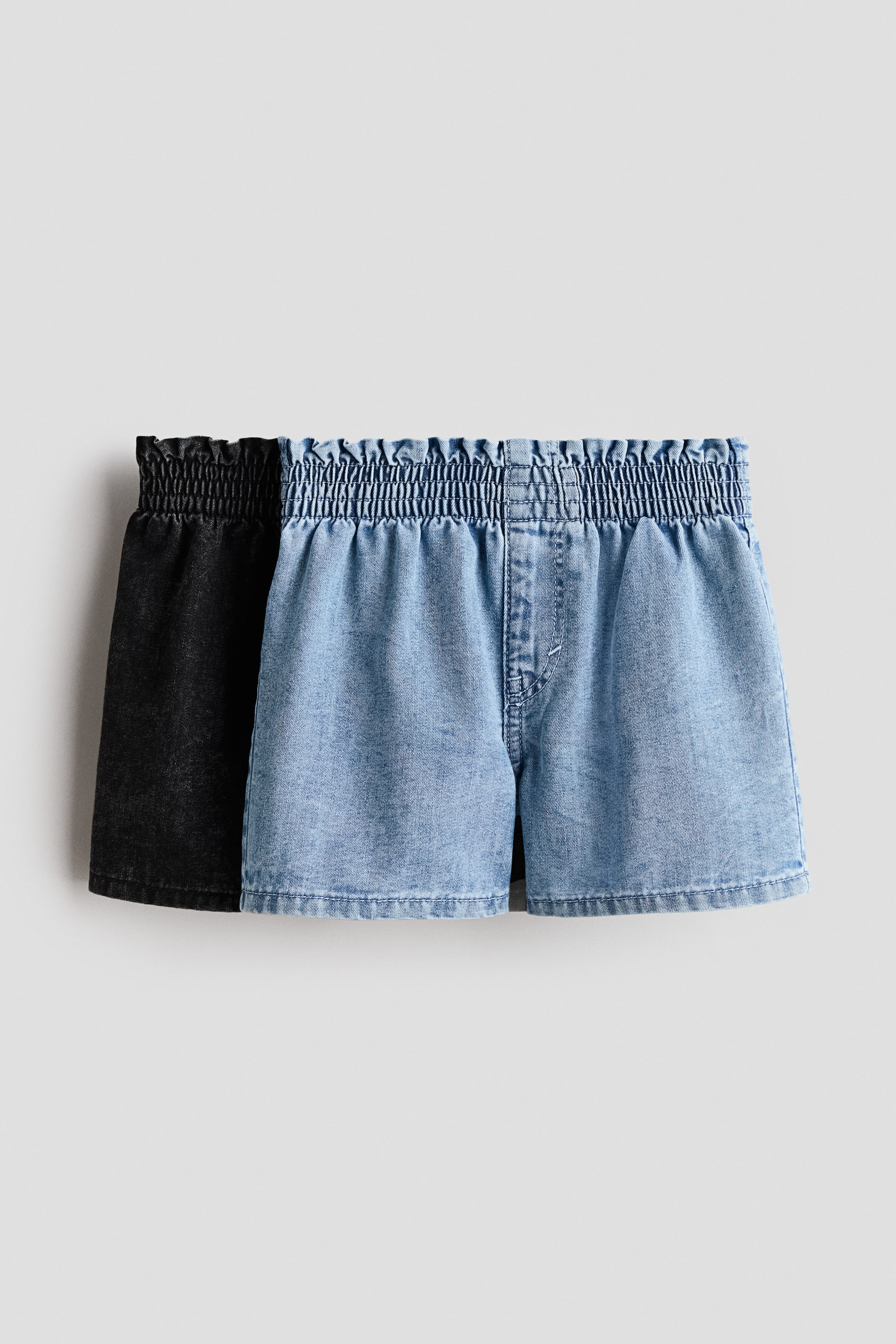 View larger image: Showcasing two pairs of loose-fitting shorts crafted from washed lightweight denim, the image presents a front view where the light blue pair slightly overlaps the black pair. Both shorts boast a paper bag waist with smocked elastication, while the light blue pair features a faux fly on the front.