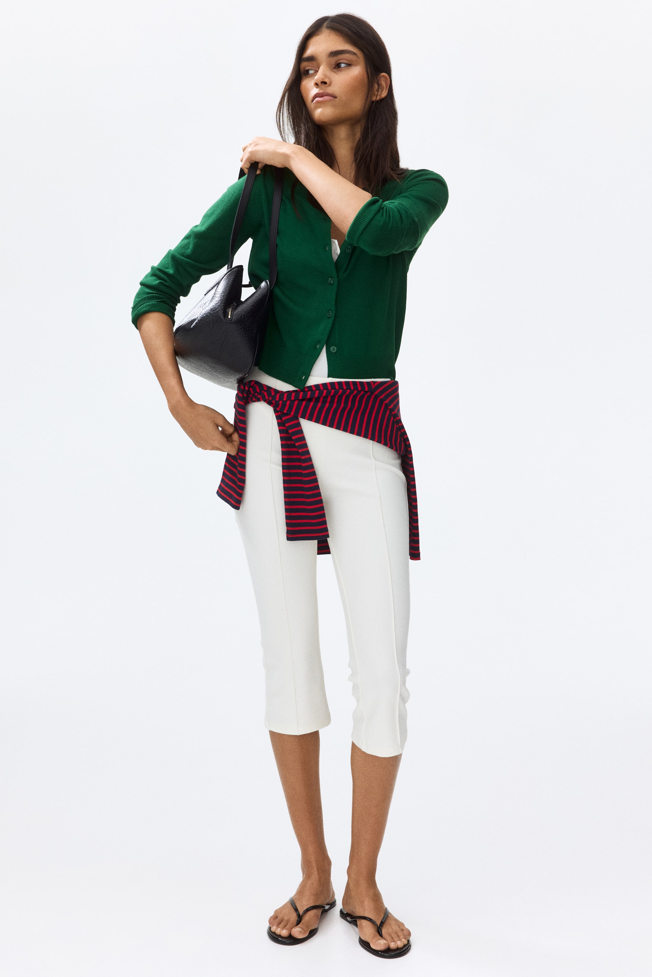 View larger image: The model is wearing a green, cropped, fine-knit cardigan that features a round, rib-trimmed neckline, buttons down the front, long sleeves, and ribbing at the cuffs and hem. It is styled over a white top, with a black handbag elegantly resting on the shoulder.