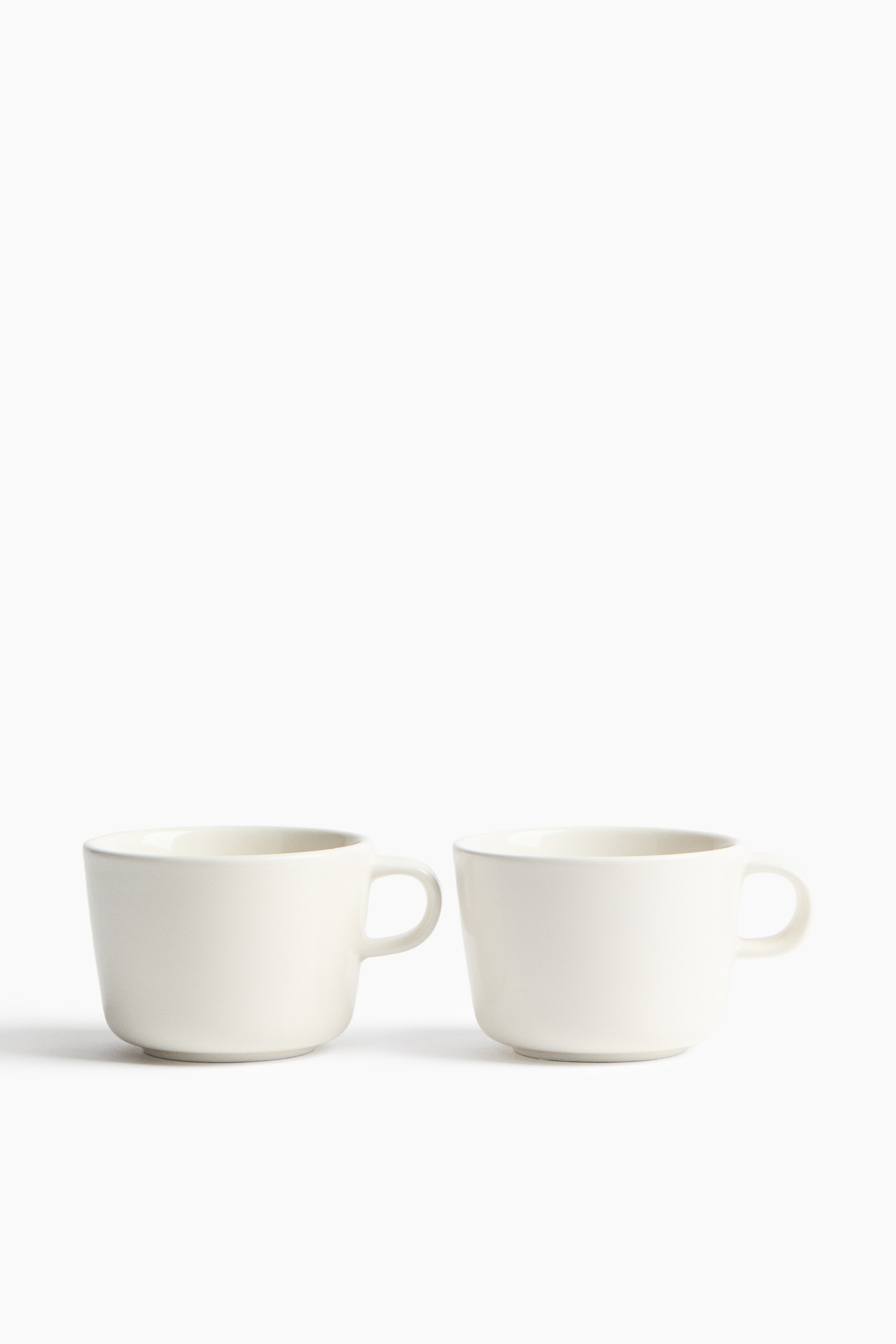 View larger image: Porcelain mugs showcase a minimalist design with a smooth, off-white finish. Two identical mugs stand side-by-side, each boasting a C-shaped handle.