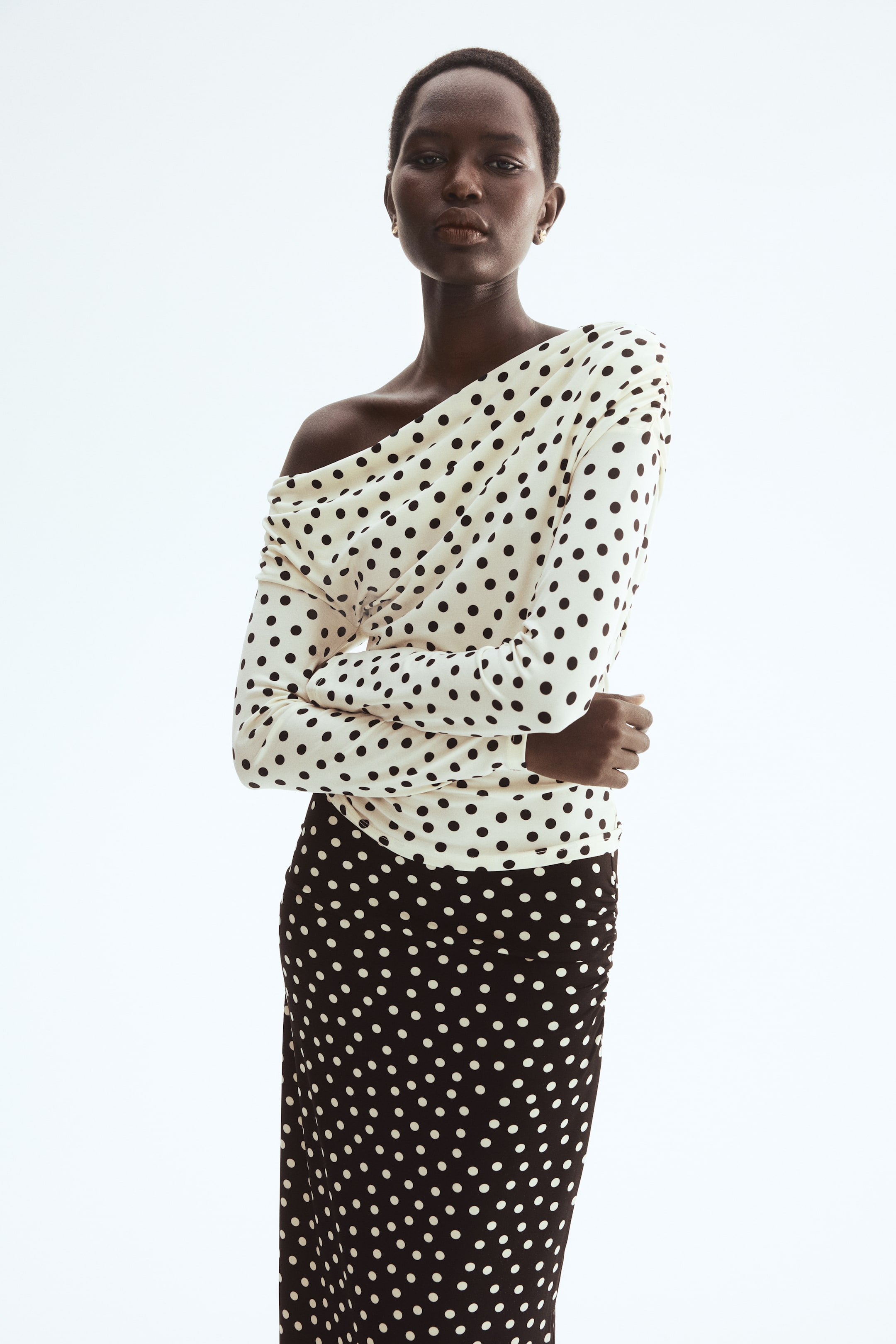 View larger image: "A cream off-the-shoulder long-sleeve top adorned with black polka dots highlights a draped neckline, perfectly paired with a sleek black midi skirt featuring cream polka dots and subtle side ruching."