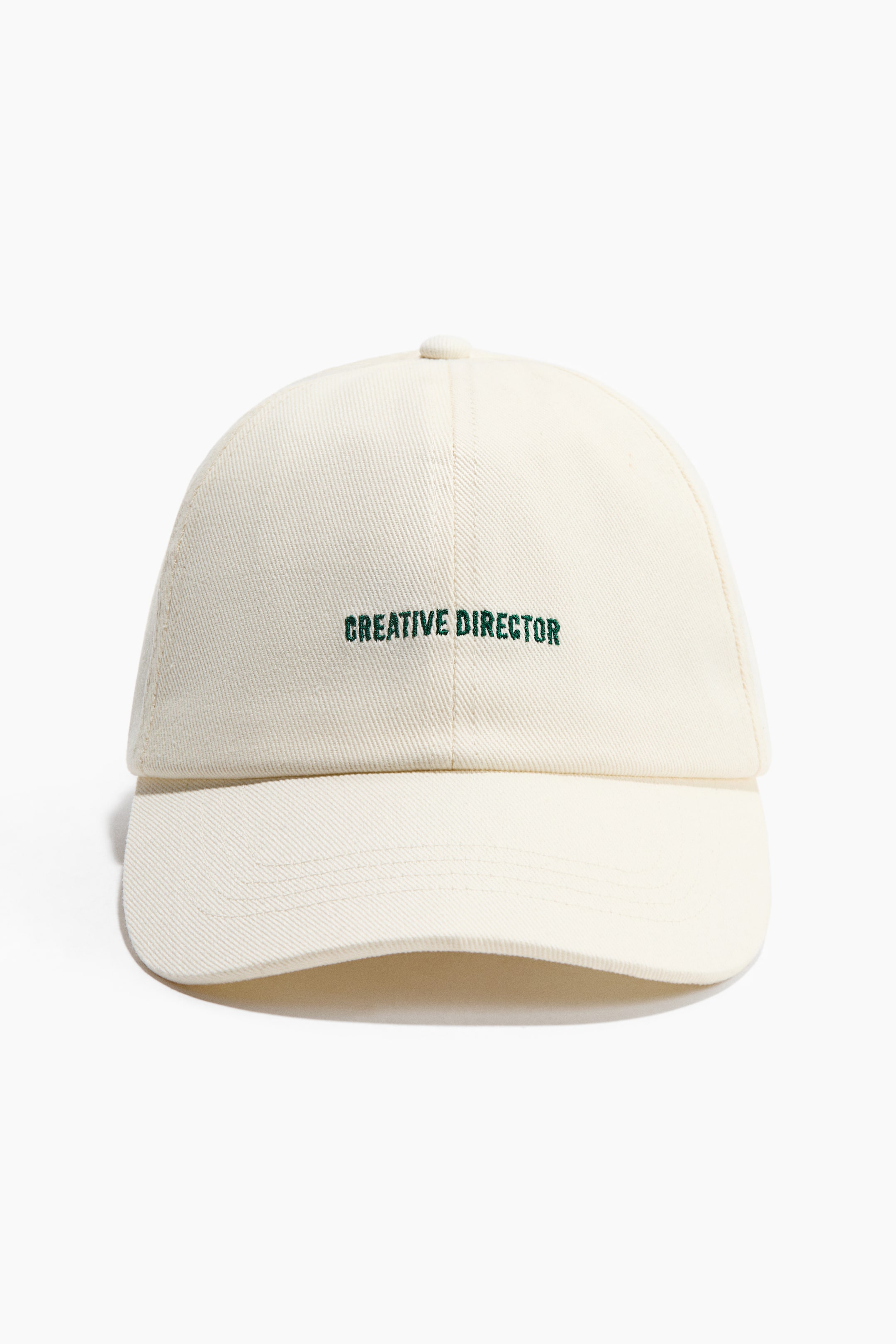 Näytä suurempi kuva: Kermanvärinen lippalakki on edessäpäin, ja sen etupaneeleihin on kirjailtu tummanvihreällä "CREATIVE DIRECTOR". Lippa on kaartuva ja ulottuu lipan pohjasta, ja vastaava nappi on lakin päällä.