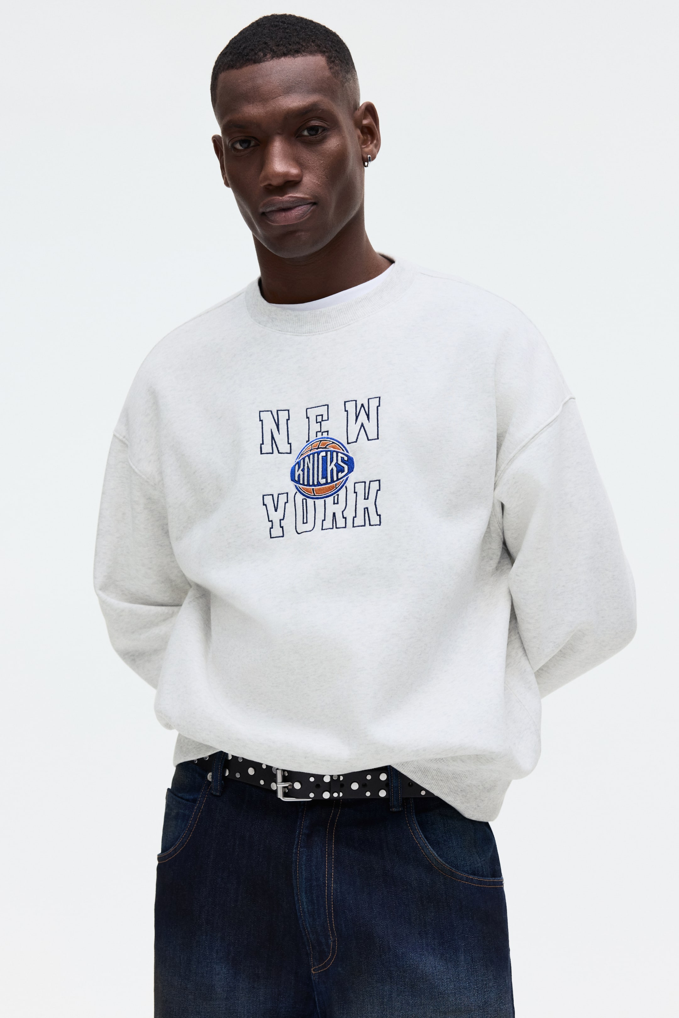 Visa större bild: En ljusgrå melerad sweatshirt med rund hals har ett broderat "NEW KNICKS YORK"-grafik med en blå och orange basketboll-logotyp på bröstet. Den har en oversized passform med ribbad rund hals och muddar, och är lätt vänd åt höger.