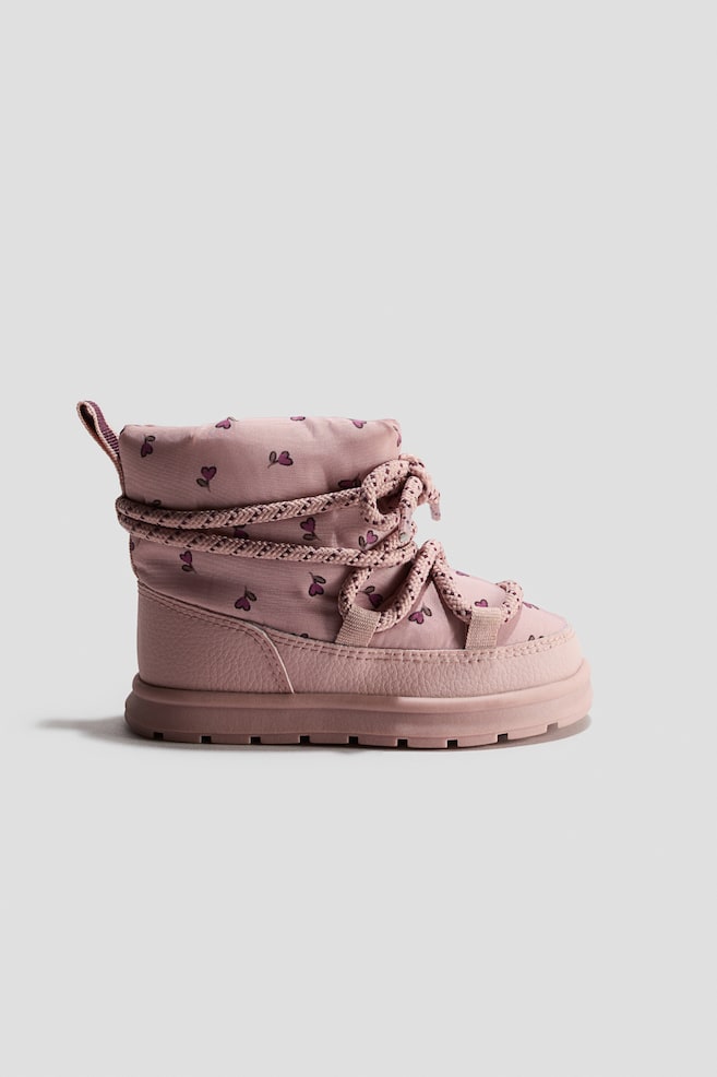 A pink children's winter boot is facing right, showcasing a padded textile upper with a purple heart print. It features thick, patterned rope laces wrapped around the ankle and a textured mudguard above a matching lugged sole. - 1