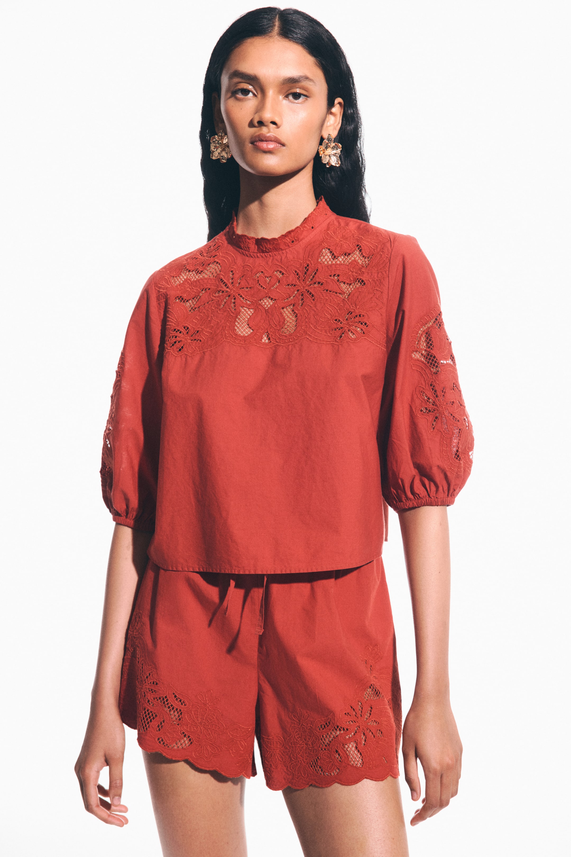 View larger image: A rust-colored matching top and shorts set is displayed. The cropped top features a ruffled mock neck, with a floral cut-out embroidered yoke covering the chest, shoulders, and elbow-length puffed sleeves, which end with elasticated cuffs. The coordinating shorts showcase a drawstring waist and a scalloped hem, embellished with matching floral cut-out embroidery.