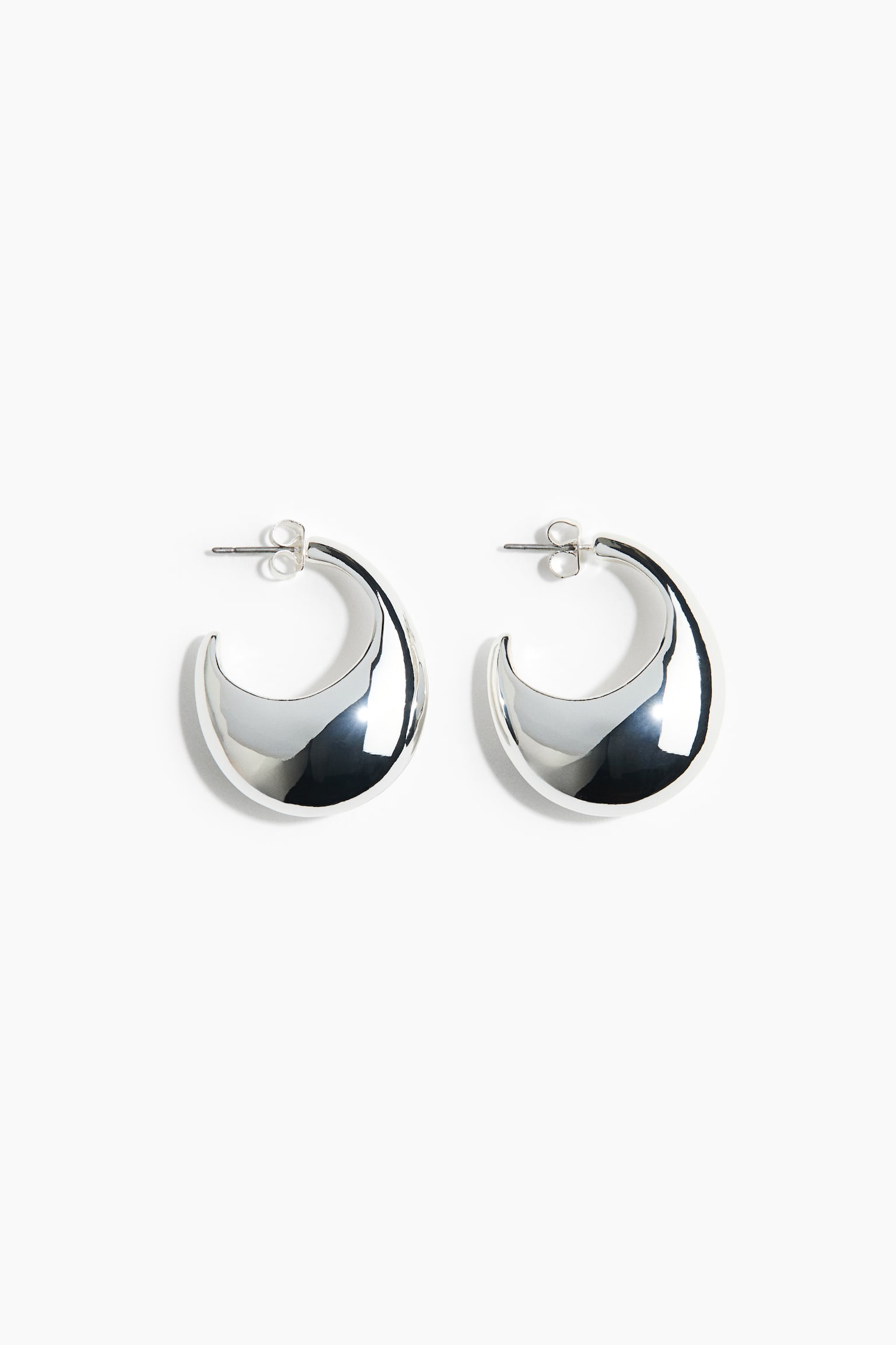 A pair of silver-toned chunky crescent hoop earrings lies flat, facing forwards. The earrings feature a highly polished, domed curved shape that tapers at the ends, with post-and-butterfly clasp backings.