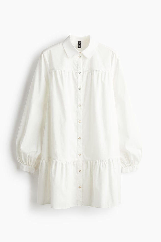 Ein weißes, langärmliges Kleid wird von vorne präsentiert, mit einem Peter-Pan-Kragen und einem vollständigen Knopfverschluss vorne. Es ist mit einer gerafften Passe, voluminösen Ärmeln mit gerafften Manschetten und einem stufigen Rock gestaltet, was eine entspannte Silhouette schafft. - 1