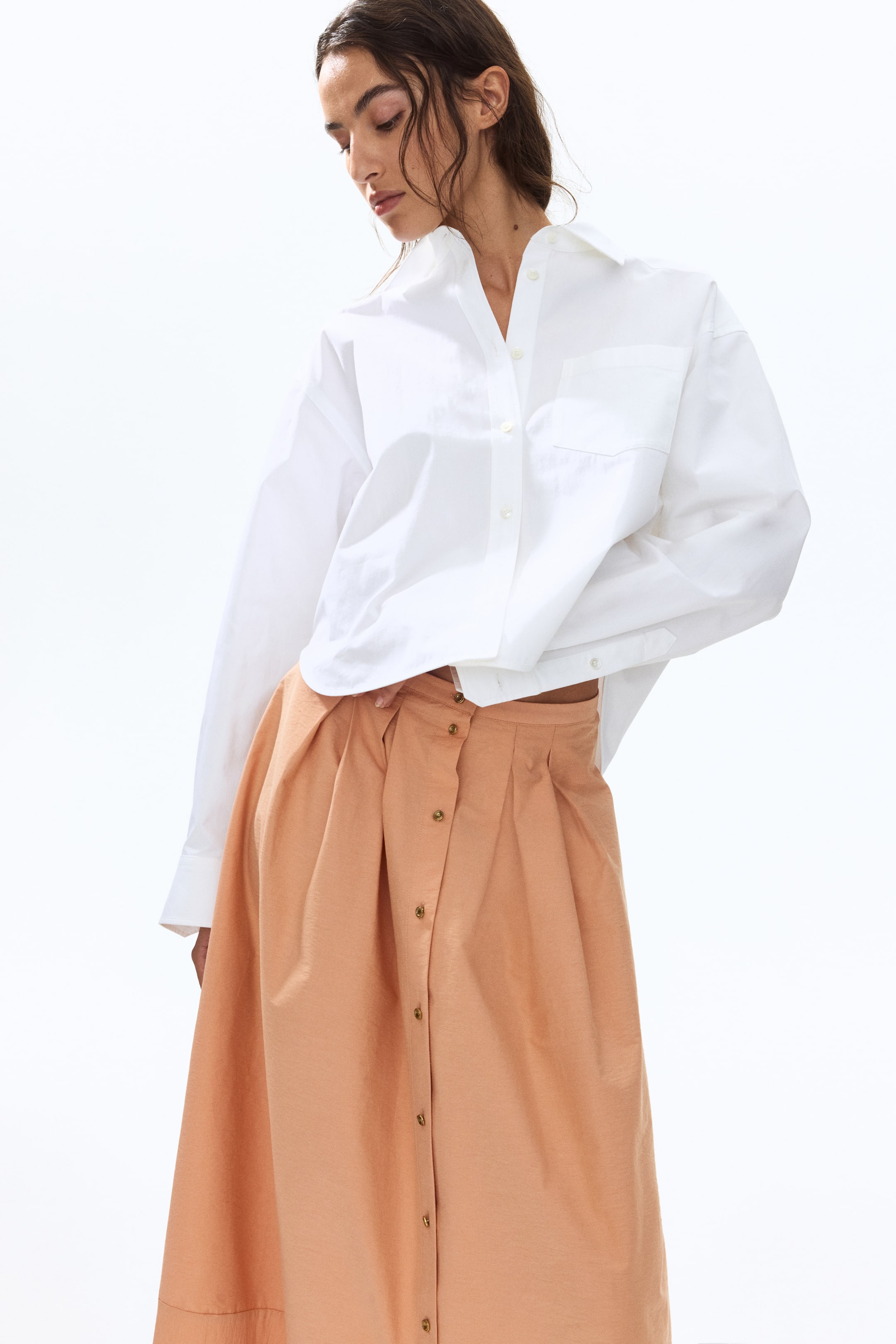 View larger image: An oversized white button-up shirt with a spread collar, buttoned cuffs, and a left chest pocket is styled loosely over a peach high-waisted midi skirt. The skirt features a full button-front closure with dark buttons and pleats.