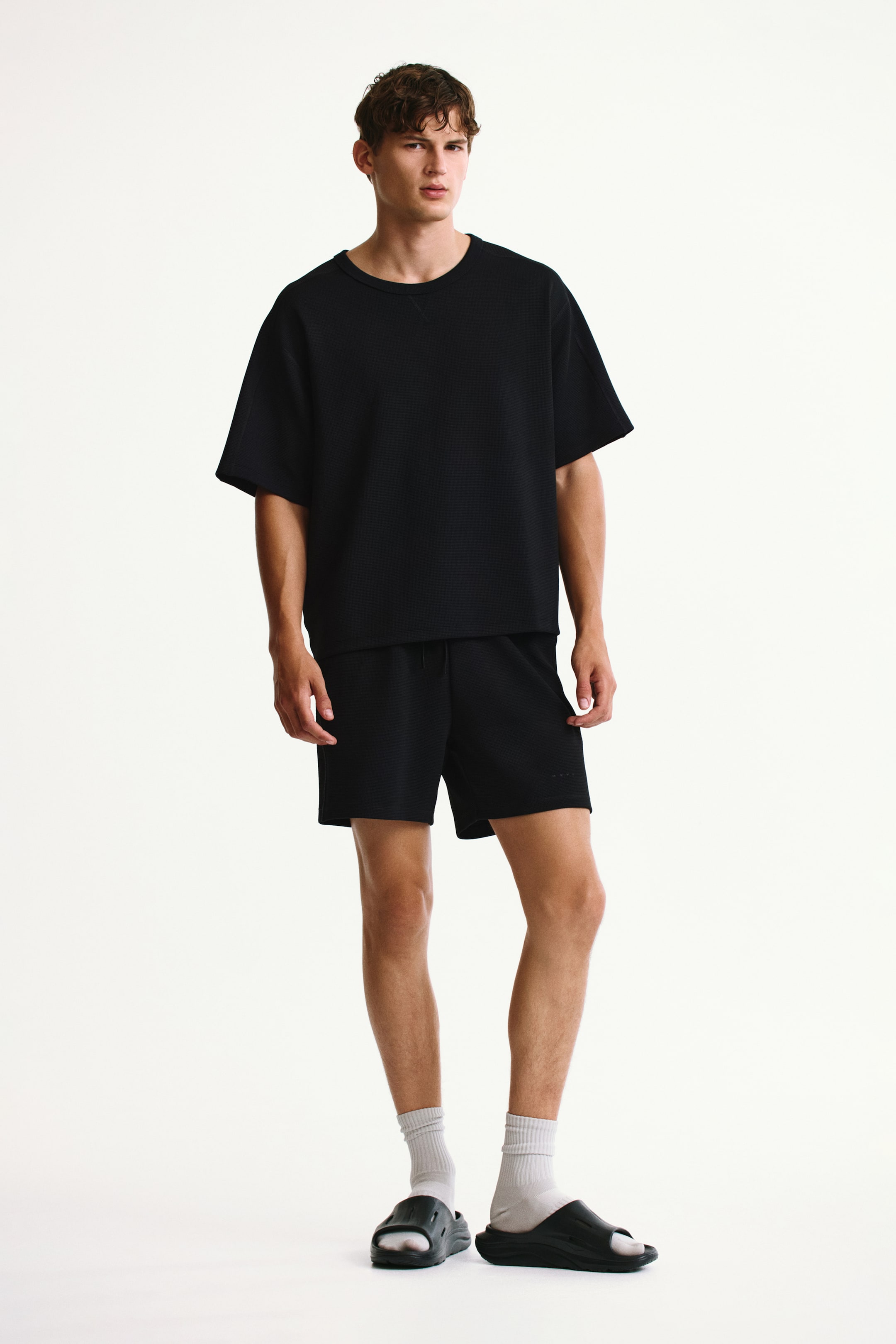 View larger image: The model is wearing black regular-fit waffled sports shorts with an elasticated waistband and drawstring. Subtle branding is featured on the lower right leg, and the shorts are styled with a matching black T-shirt, white socks, and black slides.