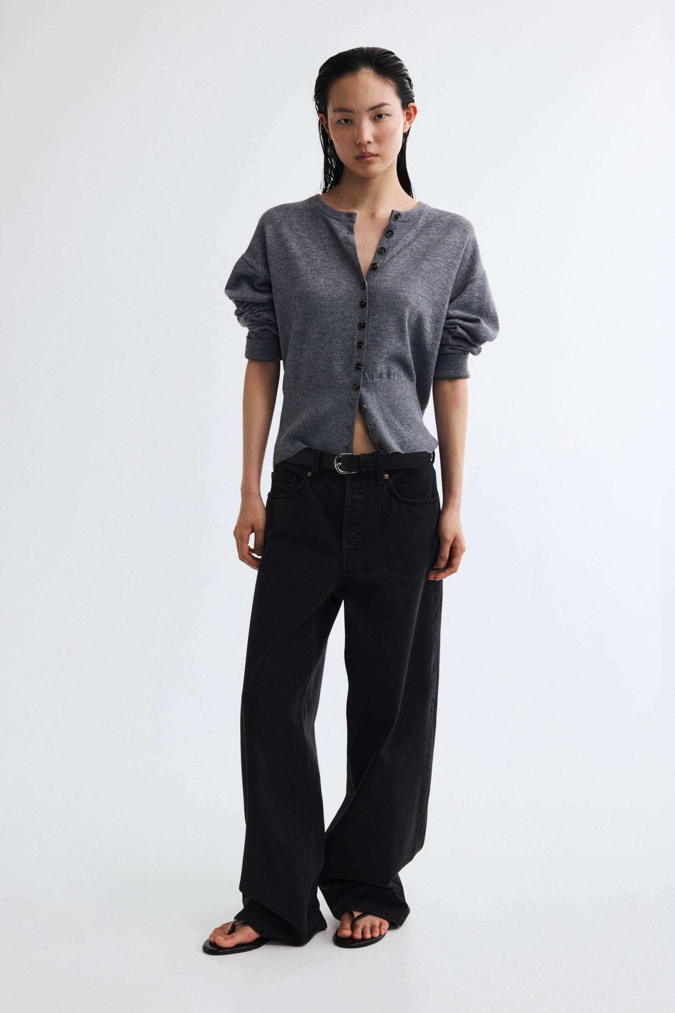 Xem ảnh lớn hơn: Người mẫu đang mặc quần jeans đen 5 túi, với cạp quần vừa phải, khóa cài nút lộ ra ngoài và ống đứng ôm rộng từ eo đến hông, rộng thùng thình từ đùi xuống gấu quần. Một chiếc thắt lưng đen được luồn qua các đỉa quần, và quần jeans chạm đến phần mu bàn chân.