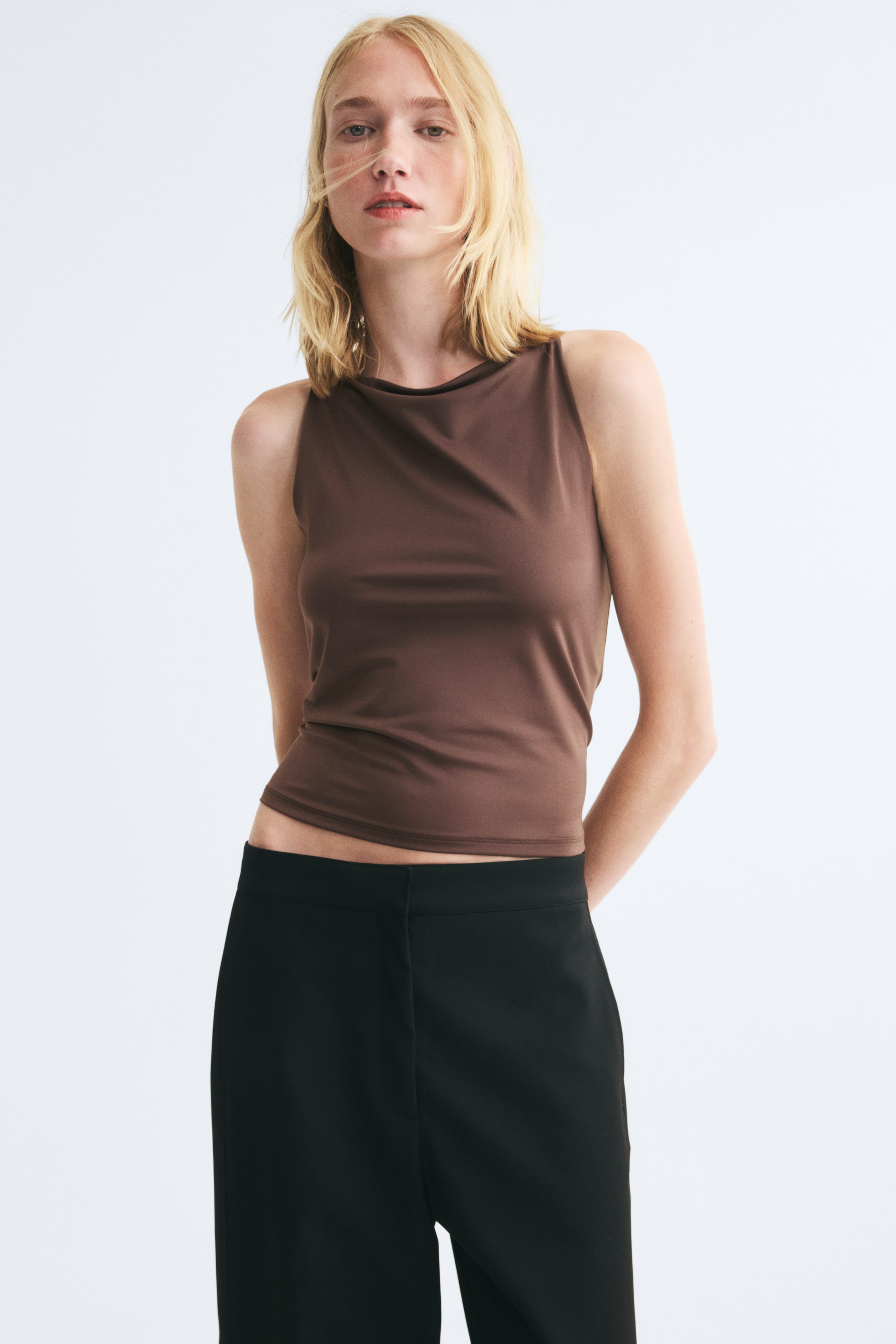 Grotere afbeelding bekijken: Het model draagt een bruin, aansluitend hemdje met een hoge, licht gedrapeerde halslijn. De top is gestyled met een zwarte broek.