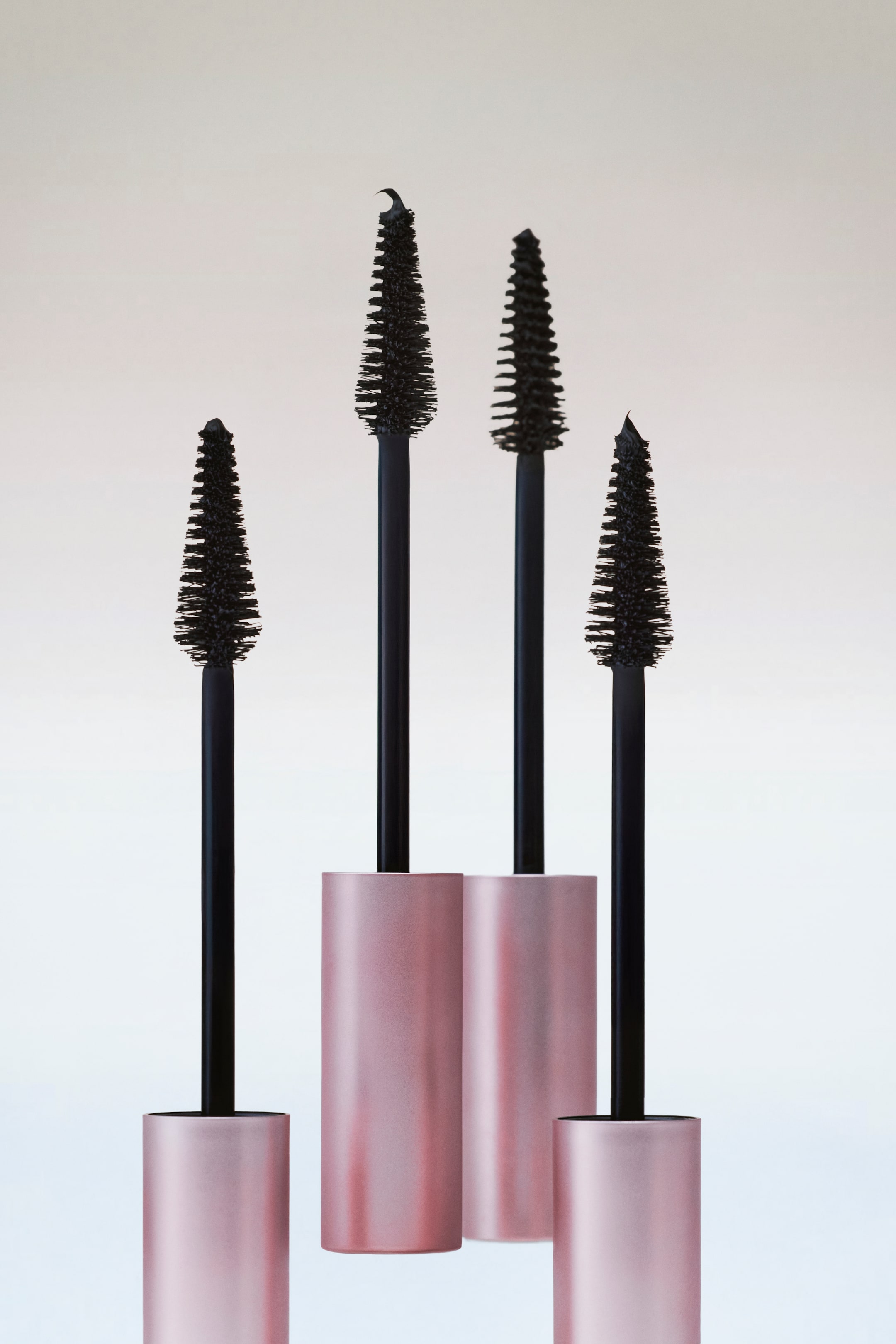 Visualizza immagine più grande: Quattro bacchette di mascara sono posizionate in verticale, ognuna con una testina a setole nere dalla forma affusolata e un gambo nero che emerge da un tubo cilindrico in metallo rosa.
