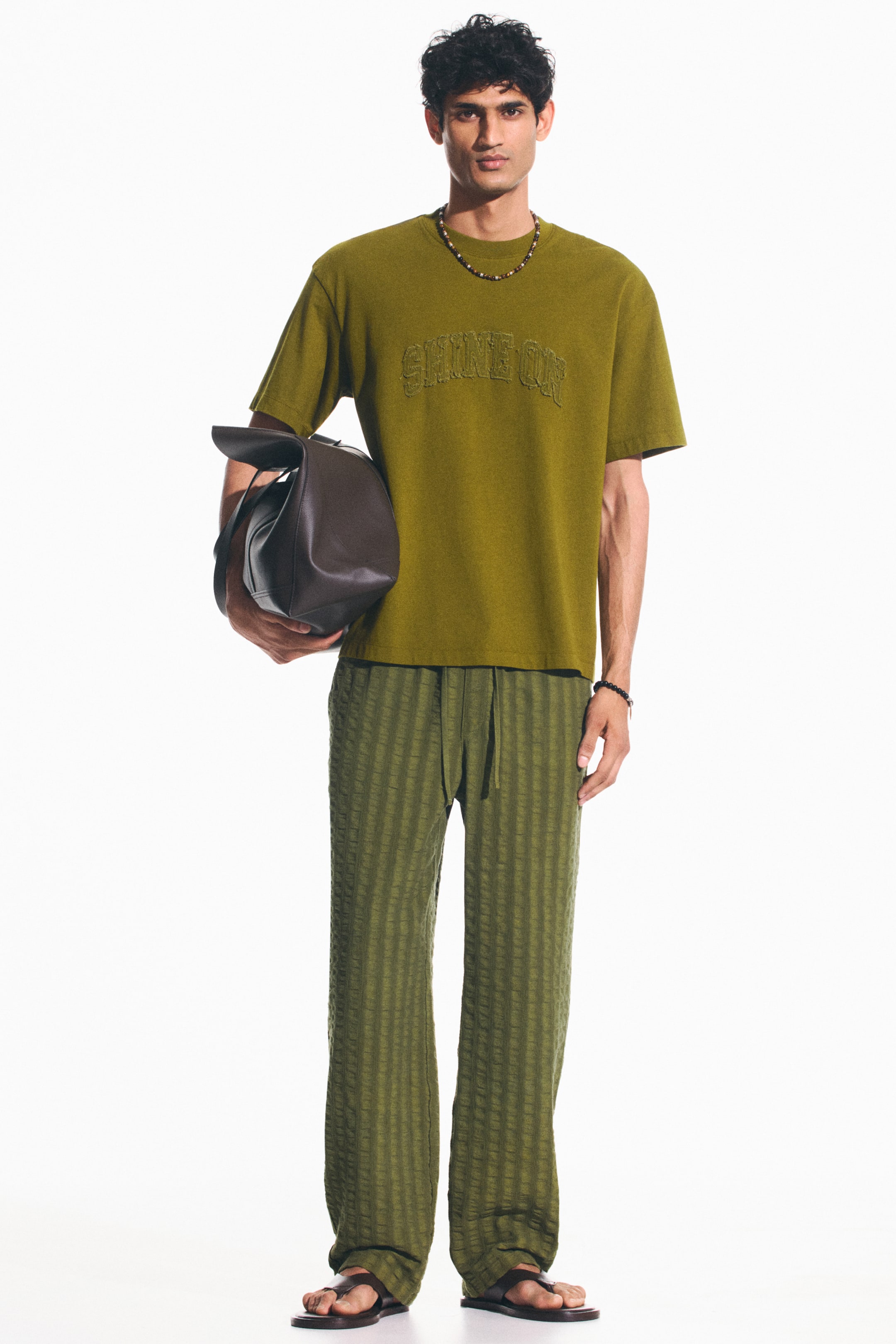View larger image: An olive green short-sleeved t-shirt, featuring an embossed "SHINE ON" text on the chest, is paired with matching olive green wide-leg trousers that showcase vertical textured stripes and a visible drawstring waist. A dark brown leather duffel bag is carried under the left arm, while dark brown leather thong sandals complete the look.