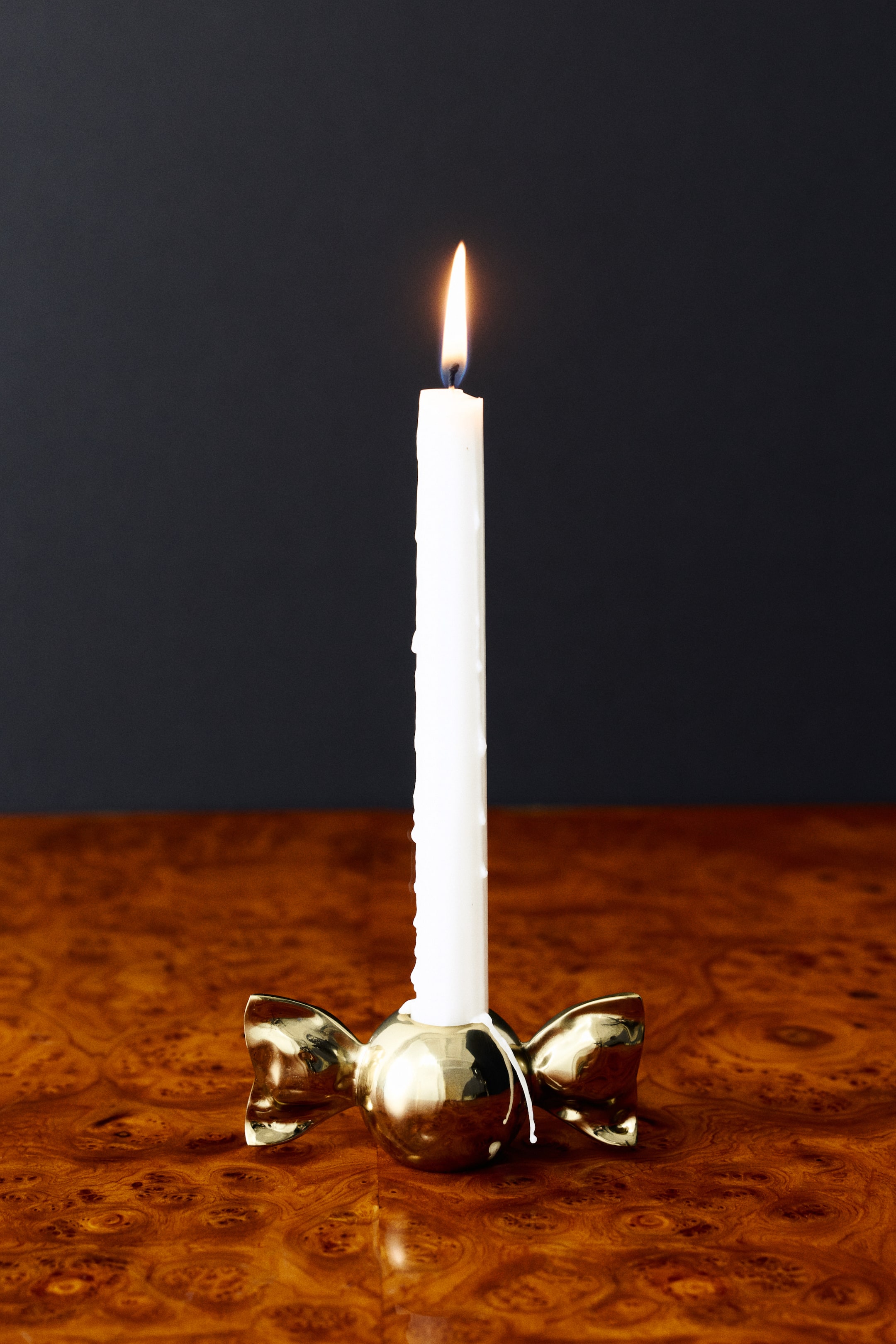 Ver imagen más grande: Un portavelas metálico con forma de caramelo, con acabado dorado pulido, sostiene una vela cónica alta y blanca con la llama encendida. El portavelas orientado hacia el frente muestra un centro esférico con extremos acampanados, similares a un lazo, que recuerdan a un caramelo envuelto.