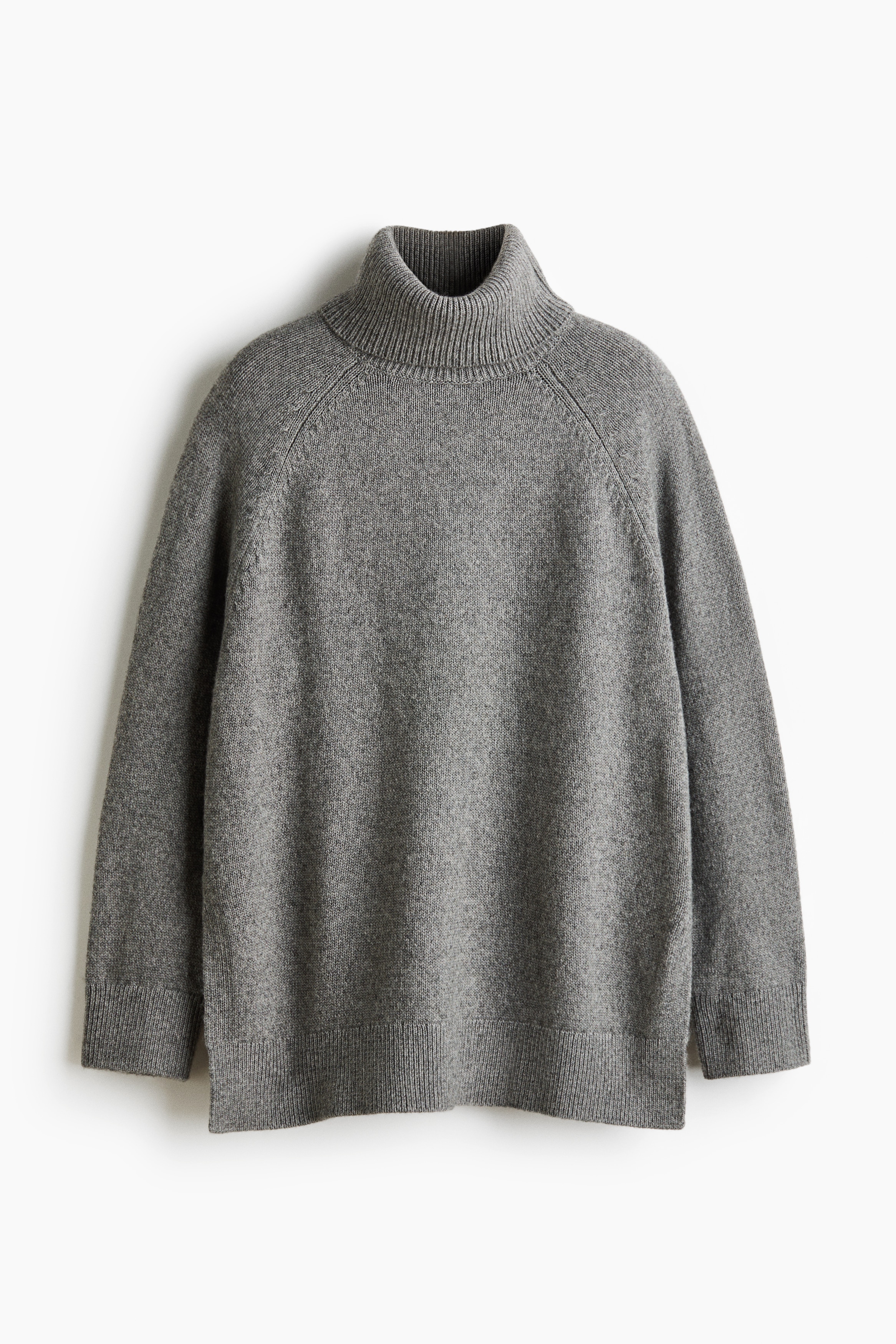 An oversized grey knit sweater featuring a ribbed turtleneck, long raglan sleeves with ribbed cuffs, and a ribbed hem with side slits is worn by the model. The model is posing with hands tucked into the pockets of cream-coloured trousers, causing the sweater’s hem to drape forward.