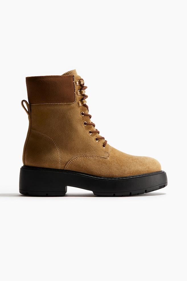 A light brown suede combat boot is presenting in a side view, featuring a dark brown padded collar, brown laces threaded through gold-toned D-rings and hooks, a thick black lug sole with a block heel, and a pull-tab loop at the back ankle. - 1