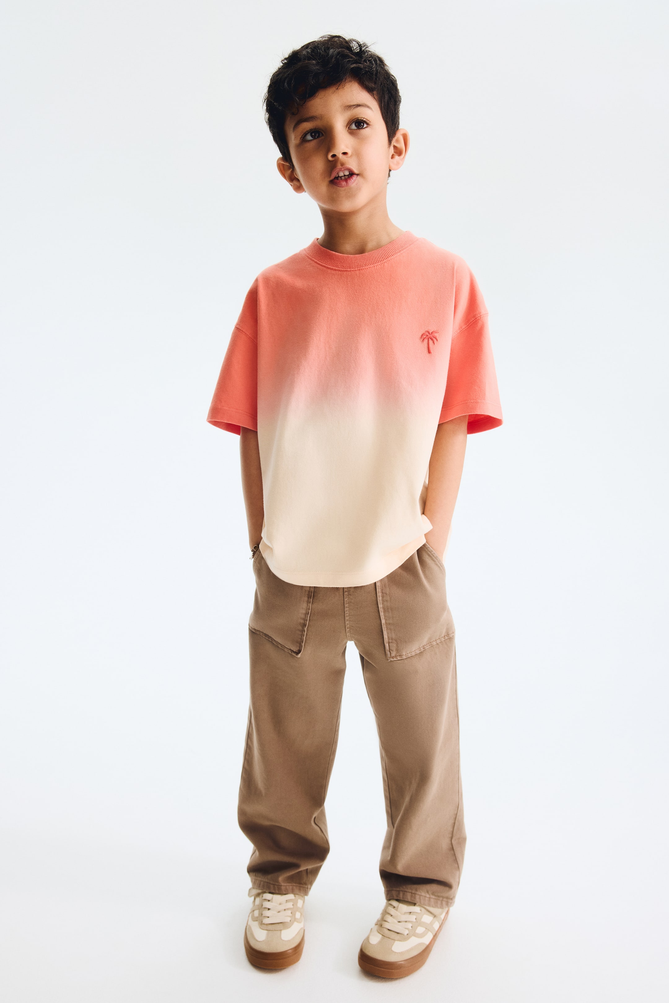 View larger image: A short-sleeve t-shirt showcases an ombré design, transitioning from coral orange at the top to light beige at the bottom, with a small embroidered palm tree on the left chest. Brownish-gray wide-leg pants complete the outfit, featuring angled patch pockets on the front and an elastic waist.