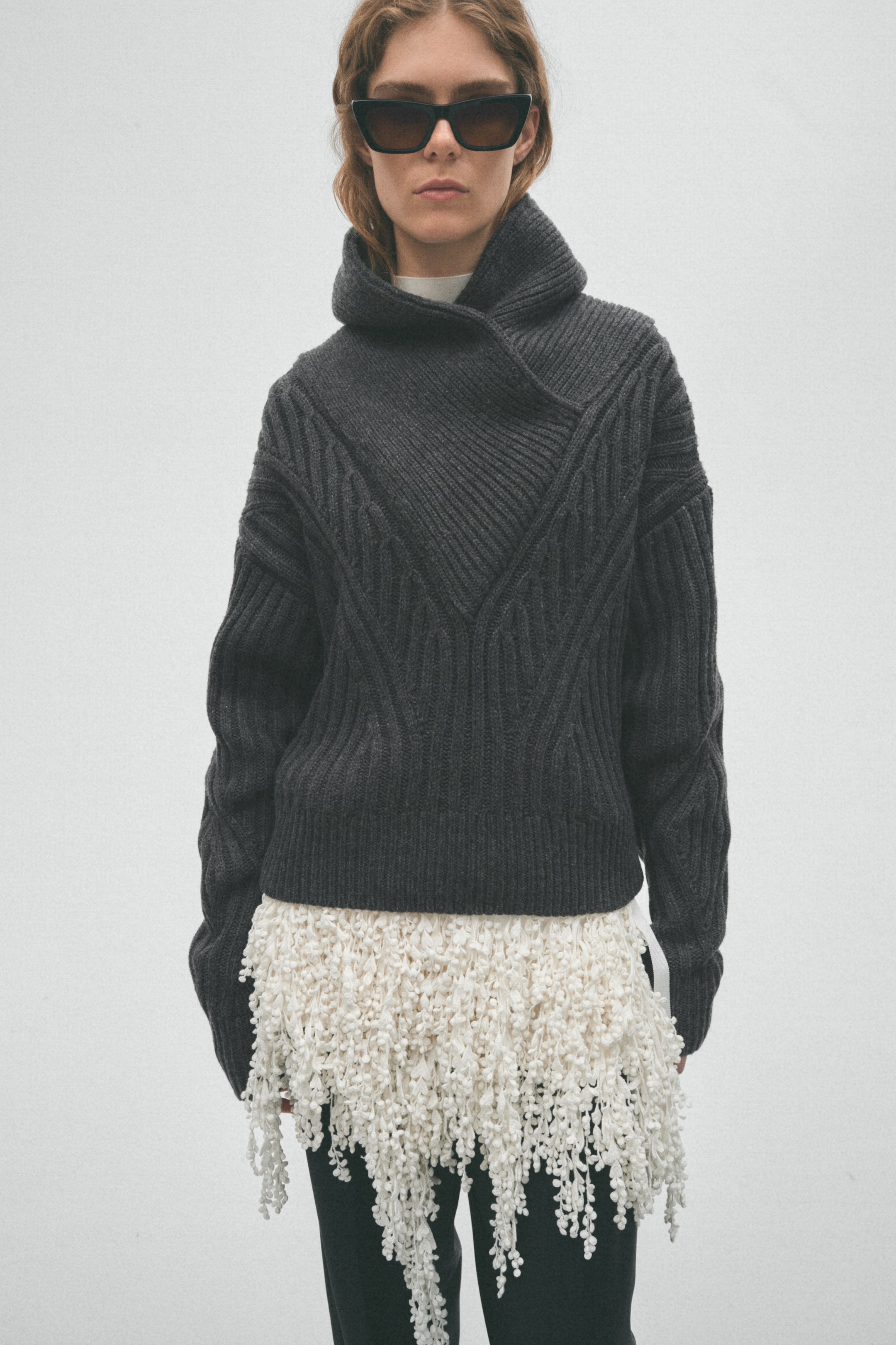 View larger image: A dark grey knitted jumper features a high, asymmetrical collar and prominent diagonal ribbed and cable knit patterns across the body and sleeves. The model is wearing the jumper, layered over a white top and a white fringed layer extending below the hem.