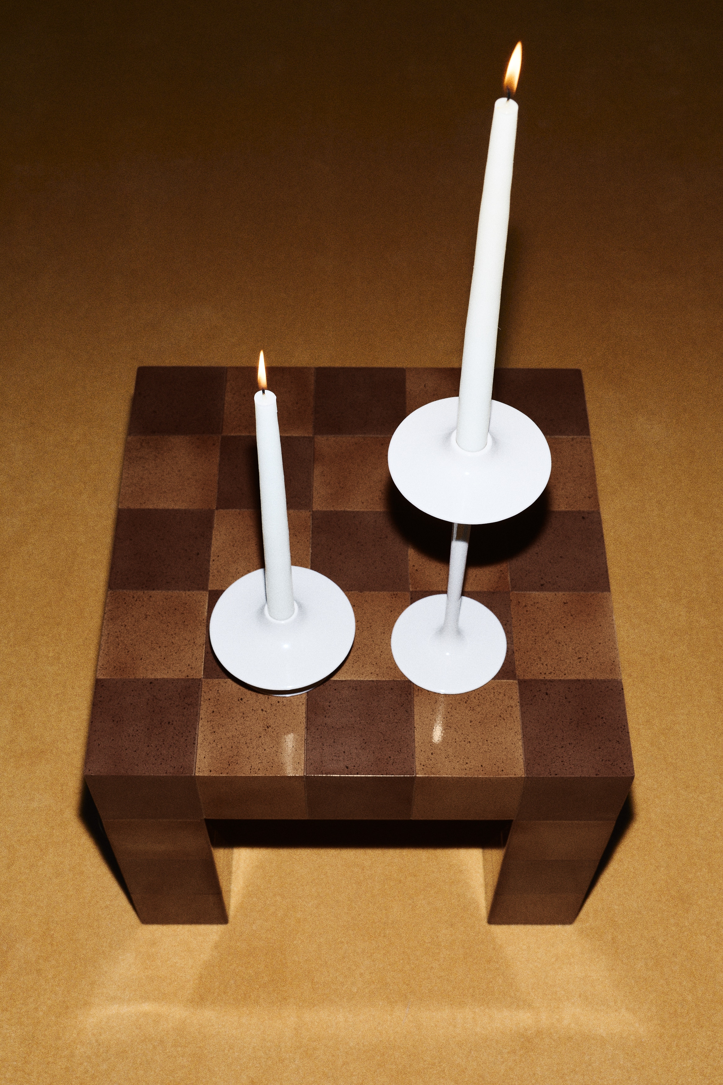A light beige candlestick is securely holding a creamy white tapered candle. The candlestick is forming a slender stem that is broadening into a flat circular dish at its top and a flat circular base at its bottom.