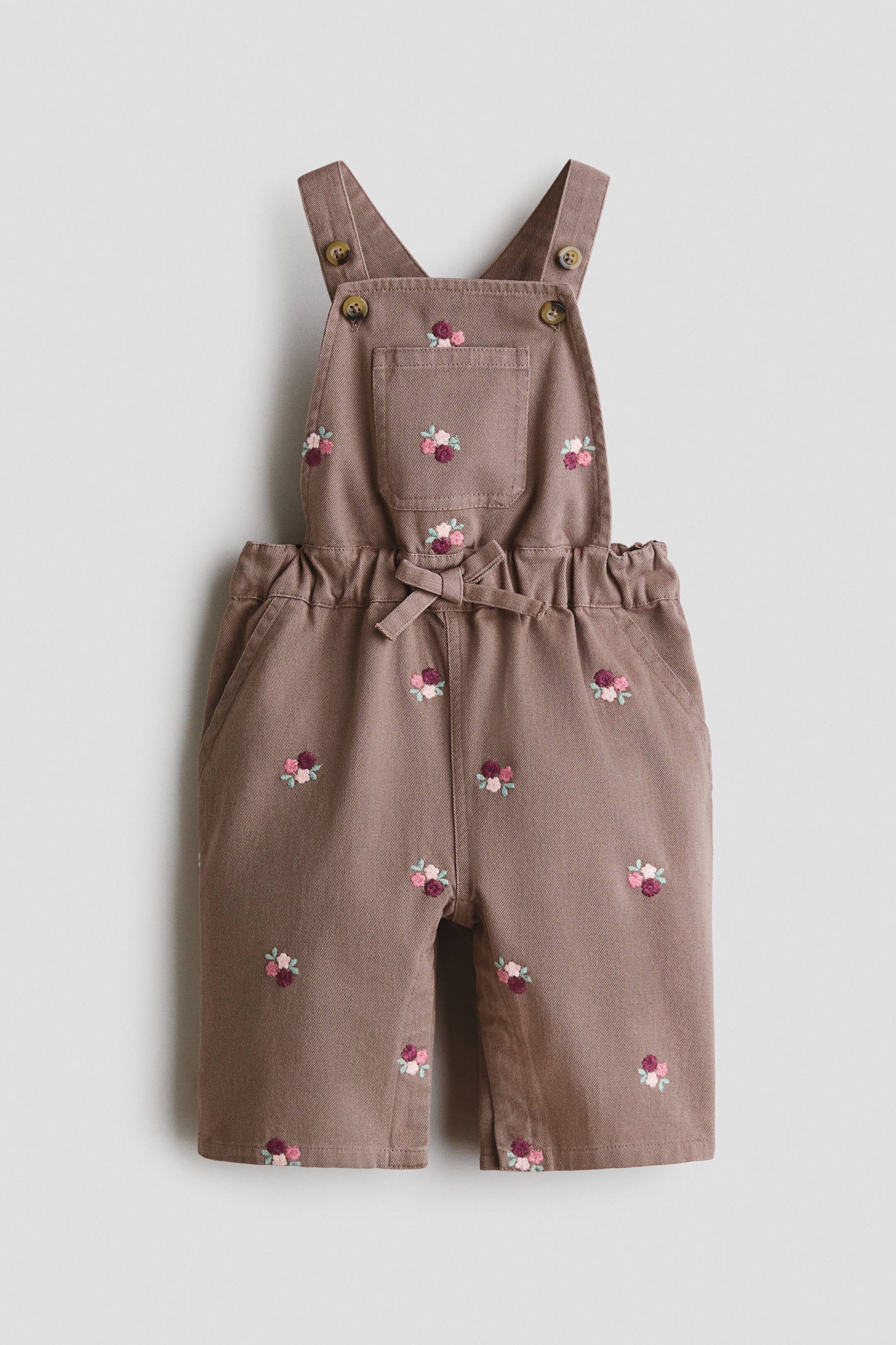 View larger image: Taupe overalls are front-facing, featuring a bib with a small patch pocket. Adjustable shoulder straps attach to the bib with brown buttons, while an elasticized waistband forms a decorative tie bow. Small embroidered floral patterns in pink, light pink, and maroon are scattered across the bib and straight-leg pants.
