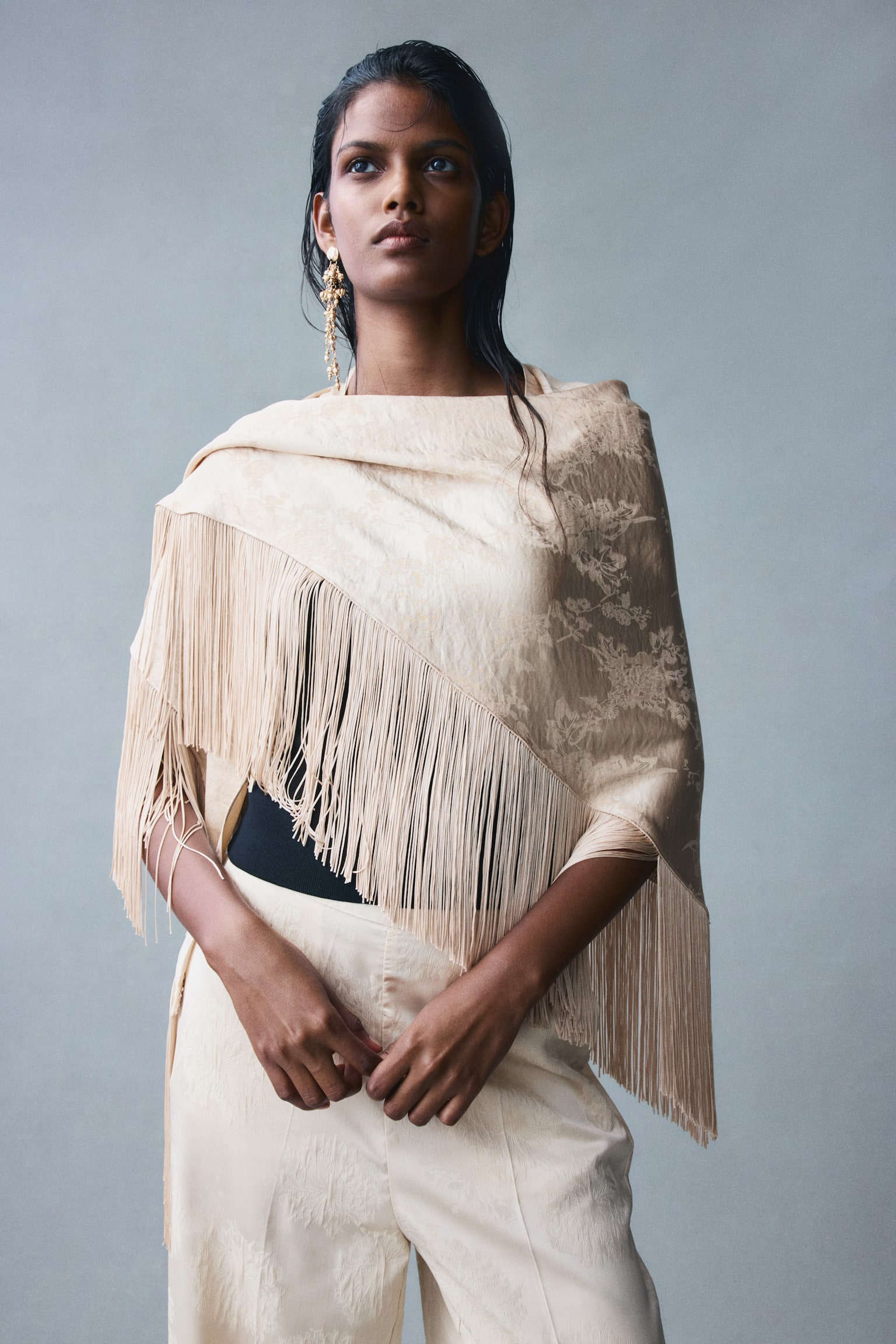 A beige open-front wrap is hanging facing the front, showing a subtle tonal floral jacquard pattern. The garment features long, matching fringe trim draping along the wide sleeves and the pointed, asymmetric hemline. - 1