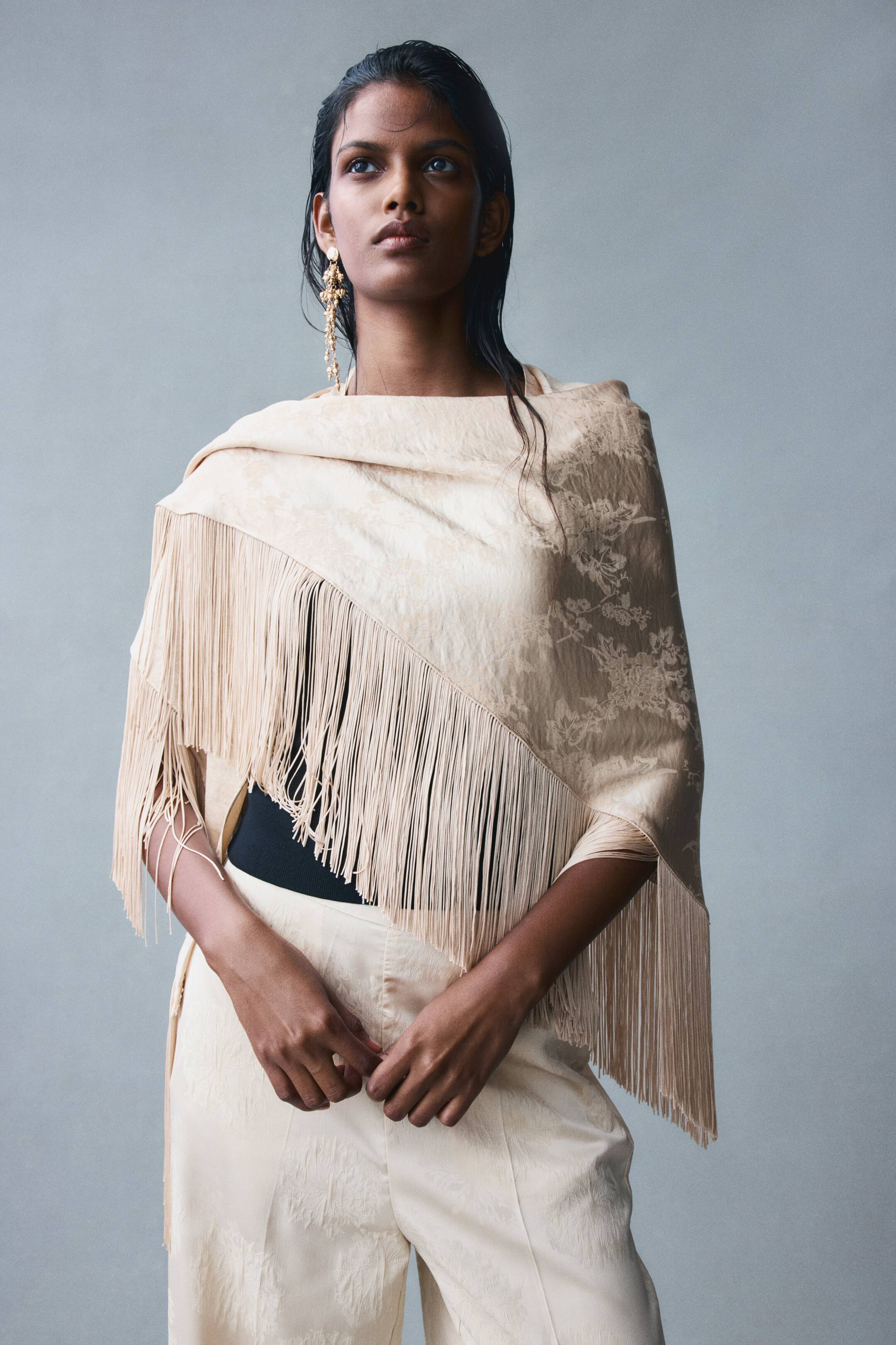 View larger image: A large, cream-colored triangle scarf in a floral jacquard weave with a subtle sheen is draped over the model's shoulders. The scarf features long, delicate fringes along the lower edges and is styled over a black top and matching cream trousers.