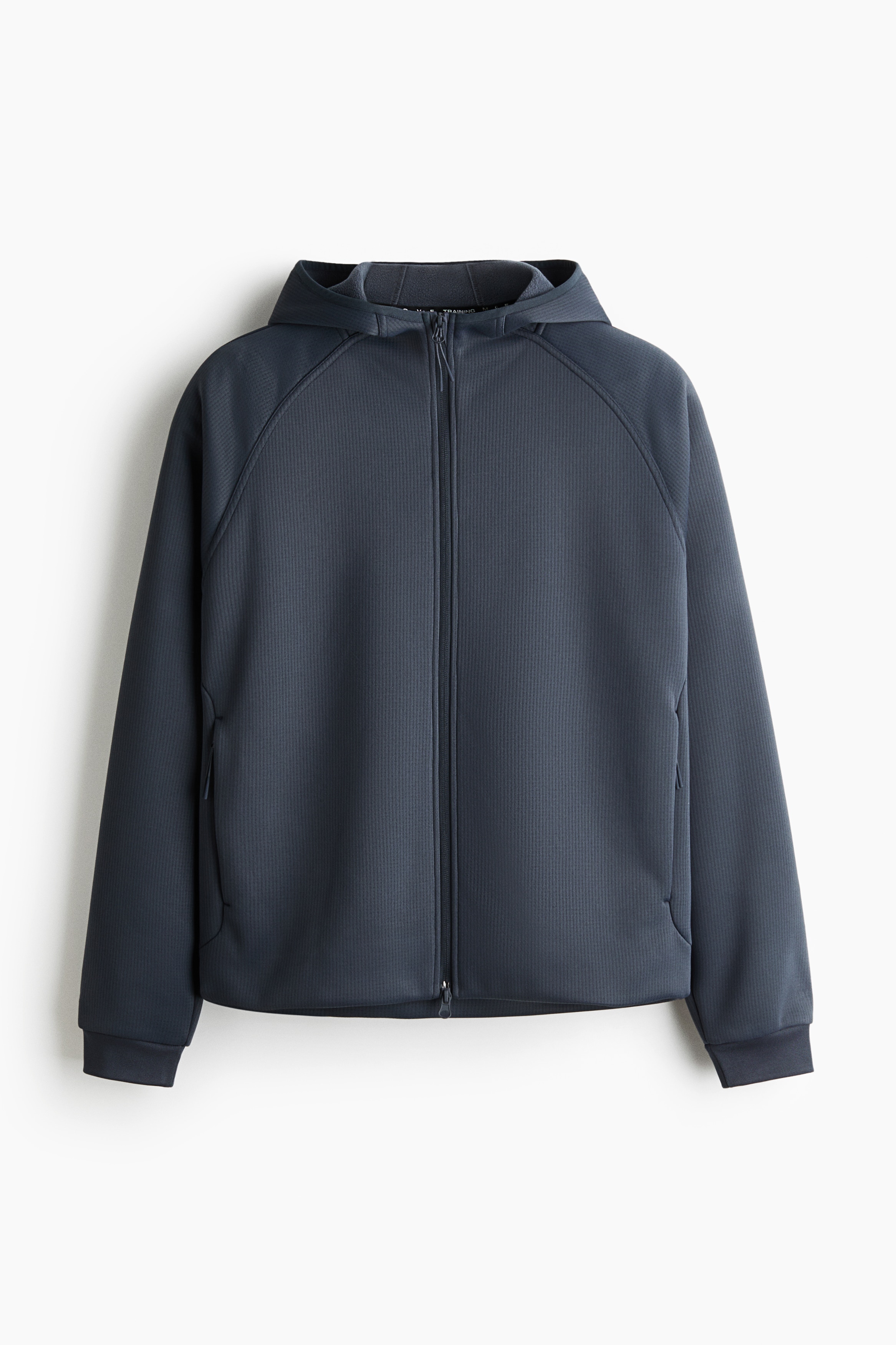A dark grey, textured zip-up hoodie is being worn by the model, featuring a front zip, a hood, and raglan sleeves. The jacket is layered over a light grey high-neck collar and styled with dark athletic shorts and grey gloves.