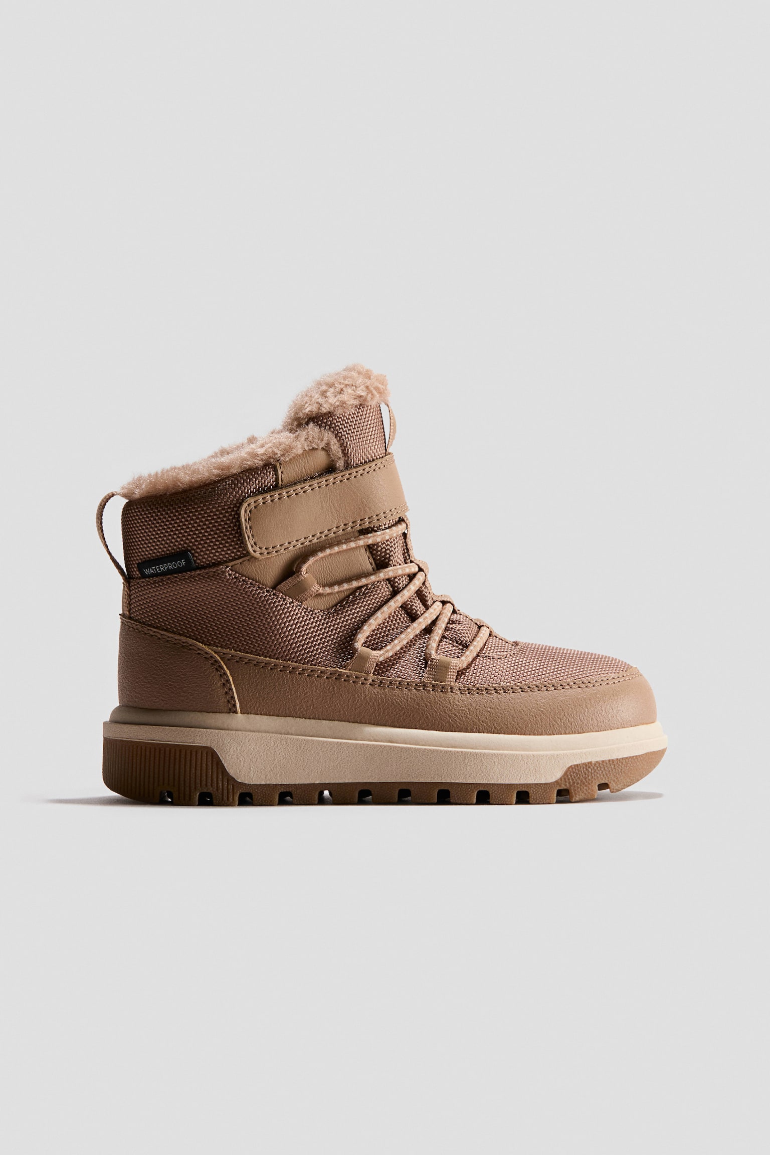 This waterproof high-top trainer is shown in side profile, featuring a tan textured fabric upper with smooth synthetic overlays and an adjustable hook-and-loop ankle strap. The shoe features criss-cross elastic laces, a plush faux-fur lining peeking out from the collar, and a thick, layered lug sole.