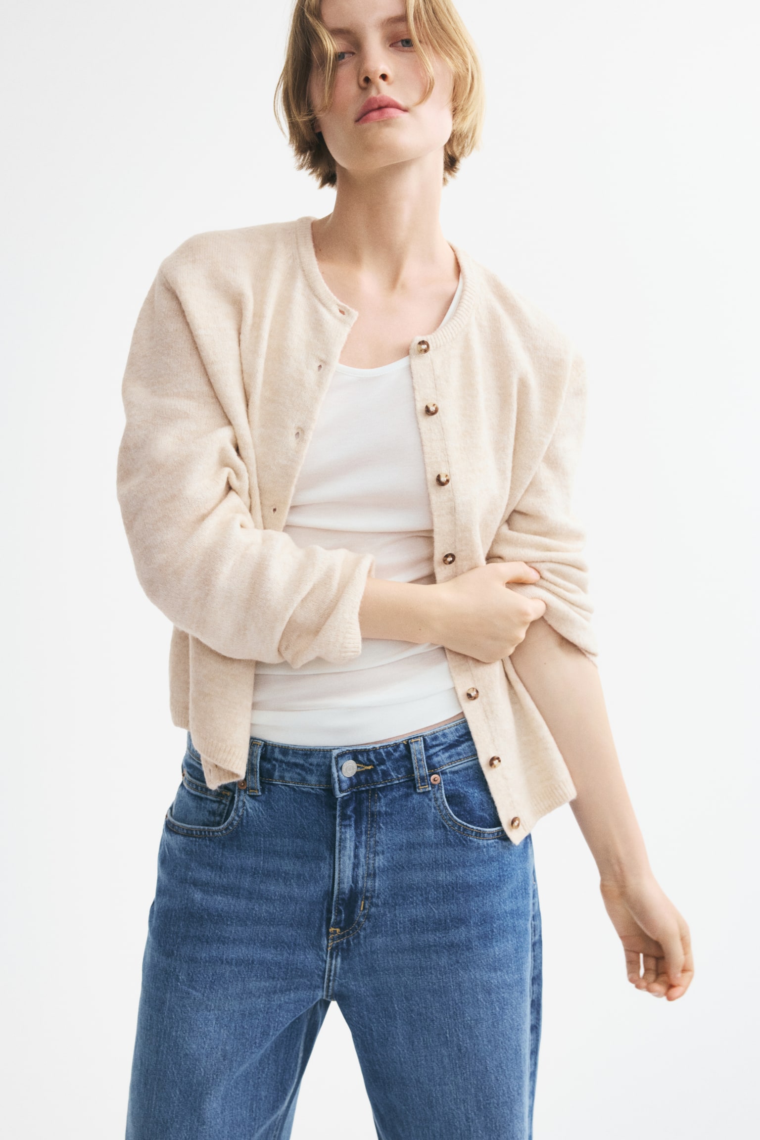 A light beige, loose-fit cardigan is showcased from the front, highlighting a soft knit texture, a round neckline with narrow ribbing, and small dark buttons running down the centre front. It features gently dropped shoulders, long sleeves with ribbed cuffs, and a ribbed hem. - 1
