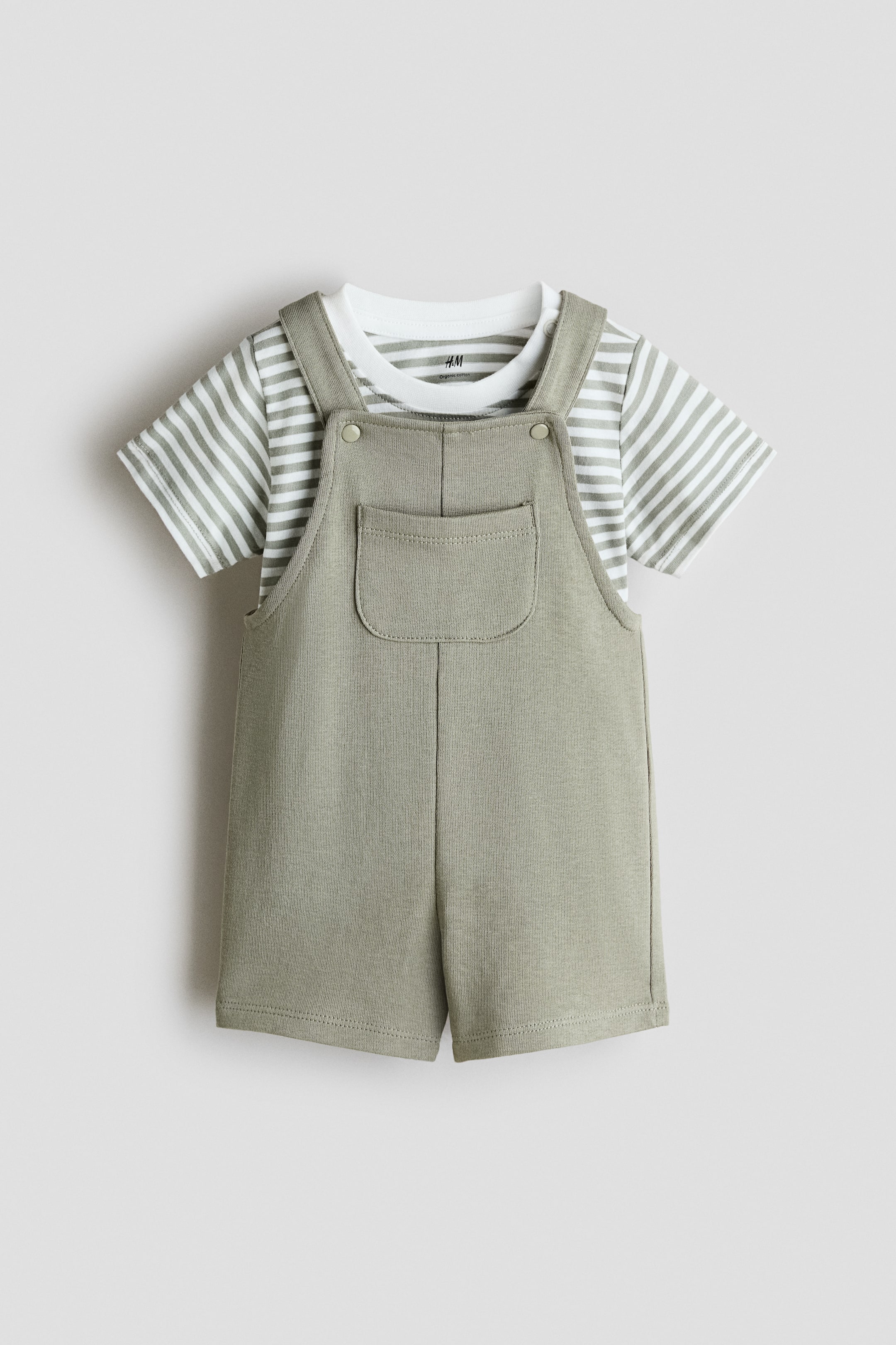 View larger image: A baby's short olive-green overall is shown from the front, with button fastenings on the shoulder straps and a patch pocket on the bib, layered over a white short-sleeved T‑shirt with light-green stripes.