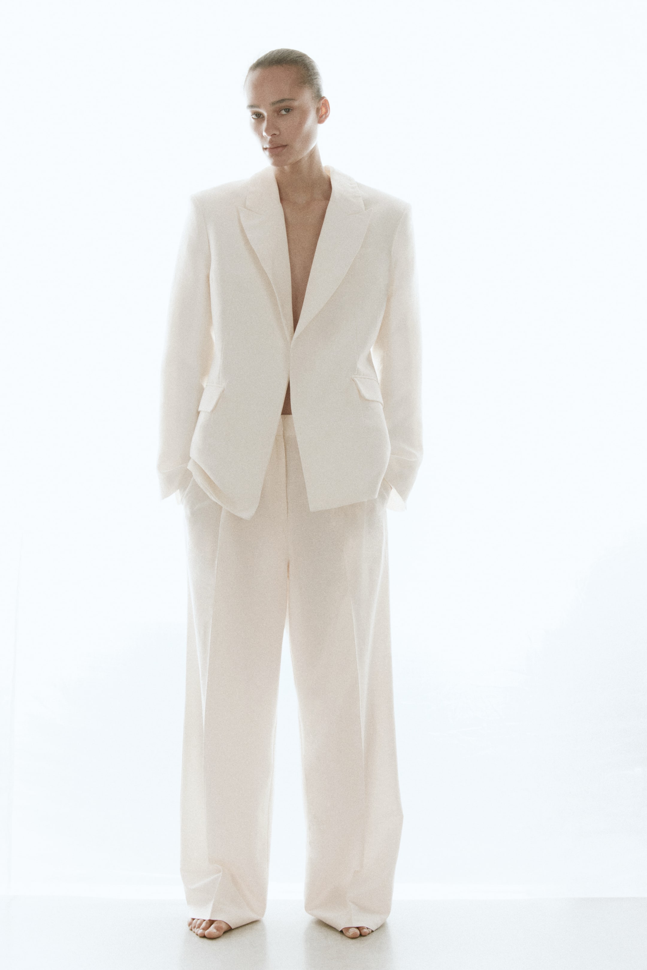 View larger image: Presenting a white pantsuit, showcasing a single-breasted blazer with notch lapels, structured shoulders, and flap pockets. The coordinating wide-leg trousers feature a pleated front and are full length.