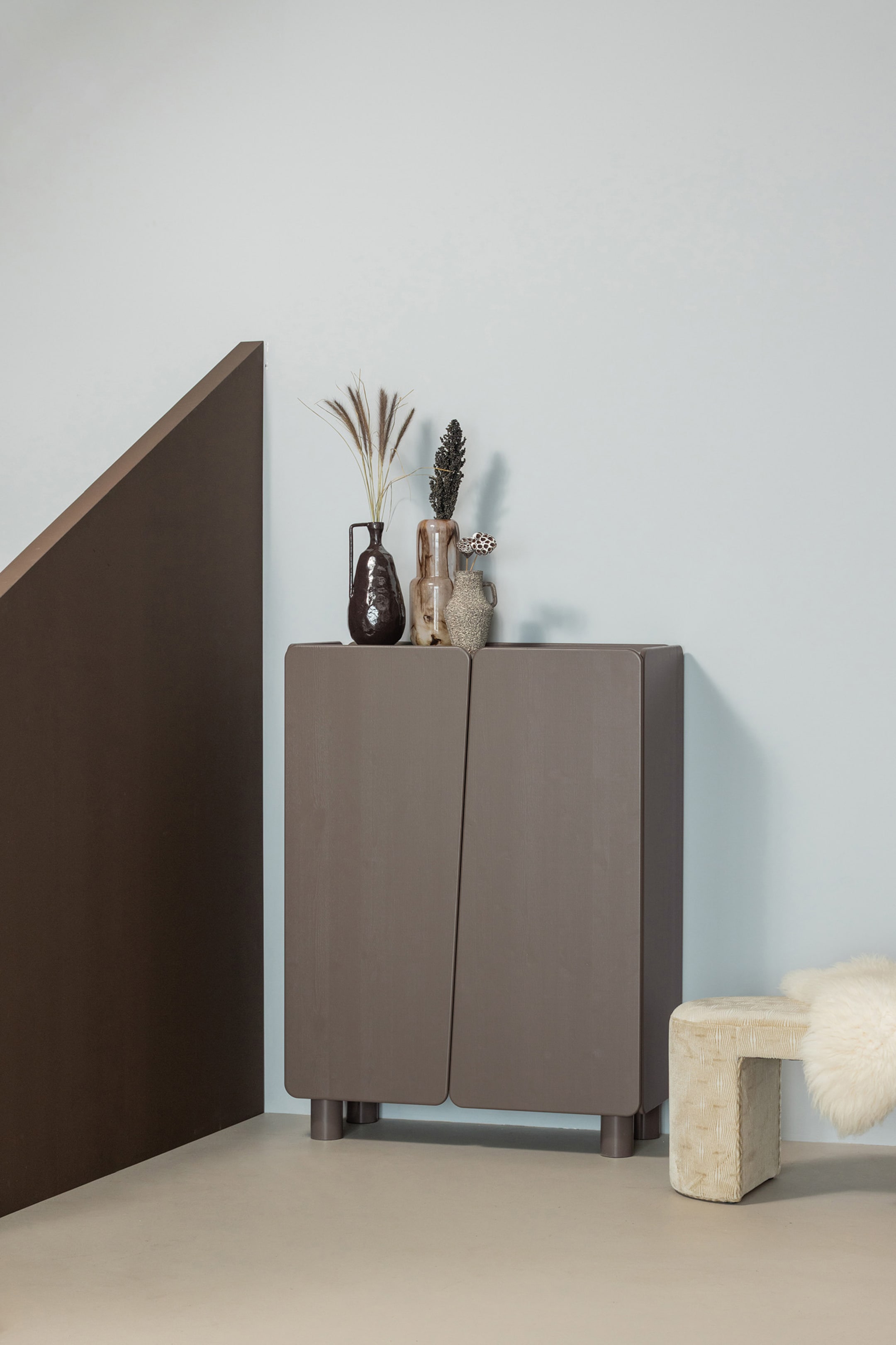 Agrandir l'image: Un meuble de couleur taupe est positionné face à l'avant, légèrement incliné vers la droite, avec deux portes présentant une couture asymétrique et inclinée ainsi que des bords arrondis. Le meuble repose sur quatre pieds cylindriques courts et sa surface supérieure plate accueille trois vases décoratifs contenant des plantes séchées.