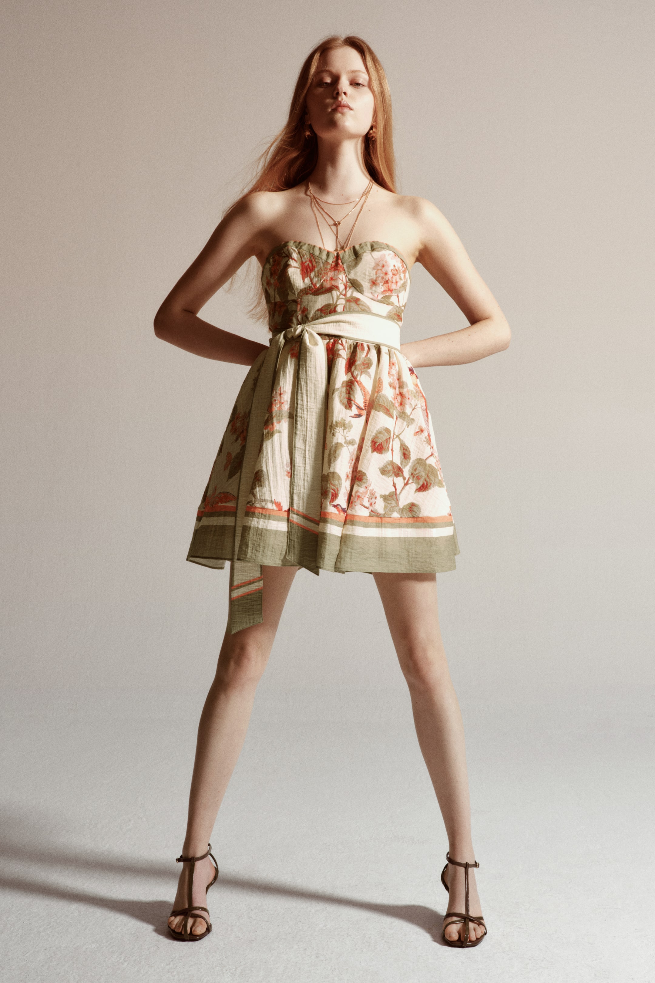 Ver imagem maior: A modelo está a usar um vestido curto tipo bandeau em tecido enrugado com padrão floral. O vestido apresenta um decote em coração, copas moldadas, um cinto atado na cintura franzida e uma saia evasé com bainha listrada, criando uma silhueta volumosa.