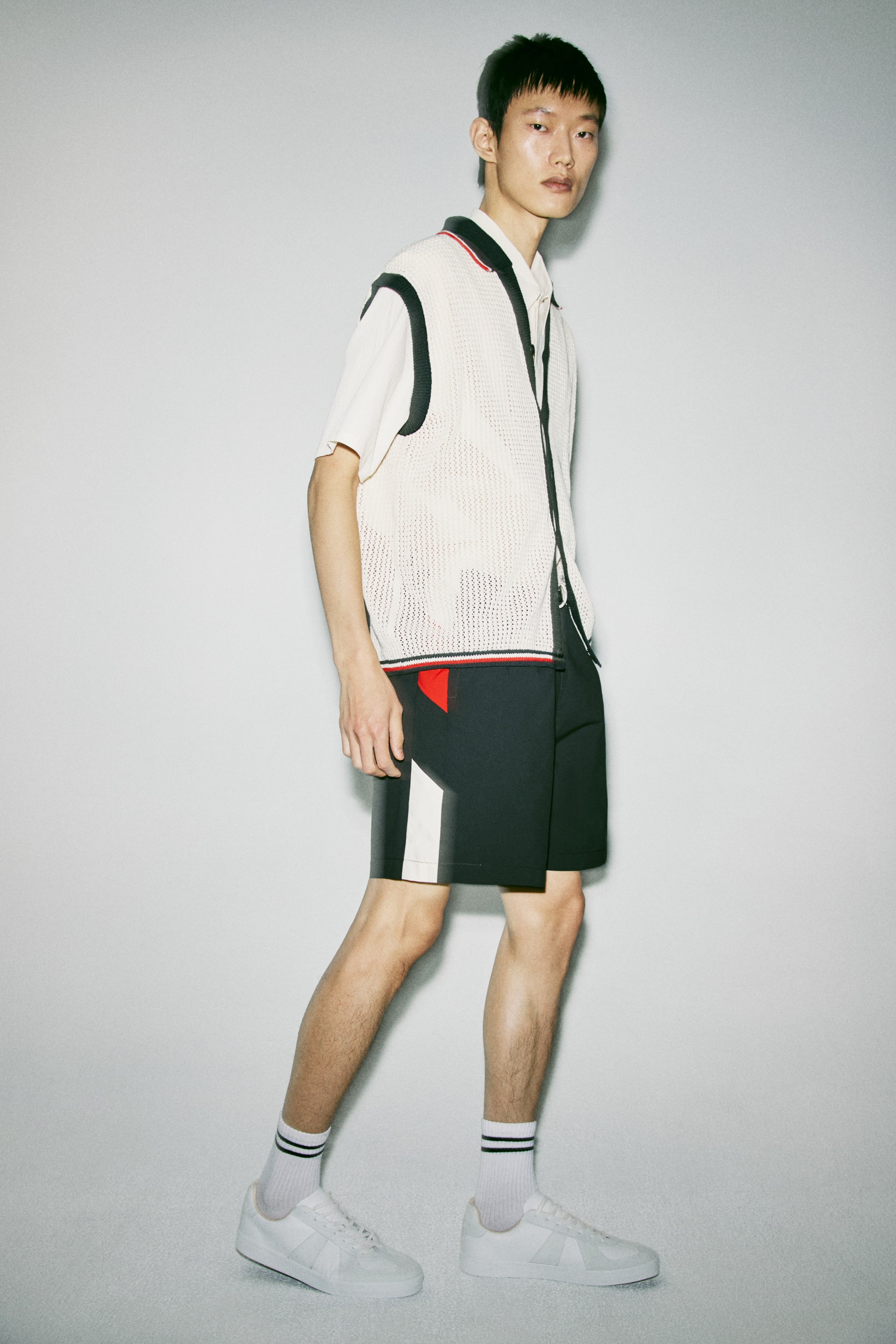 View larger image: A cream-coloured open-knit vest with black trim, a black-and-red striped polo collar and a red, white and blue striped hem layers over an off-white short-sleeved polo shirt. Black shorts complete the outfit, featuring a white geometric stripe on the left leg and a red triangular design on the right, paired with white sneakers and white crew socks with black stripes.