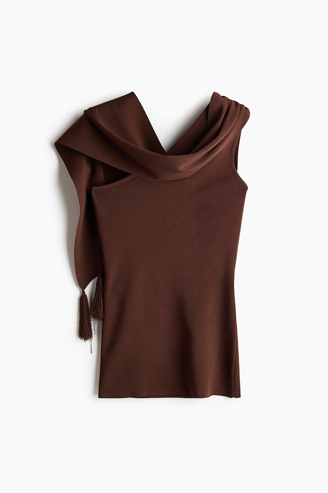 A brown fitted one-shoulder top in a soft, fine knit is featuring an asymmetric neckline and a wide shoulder strap. This strap is extending into a wide scarf detail embellished with tassels at its end, draping down the side. - 1