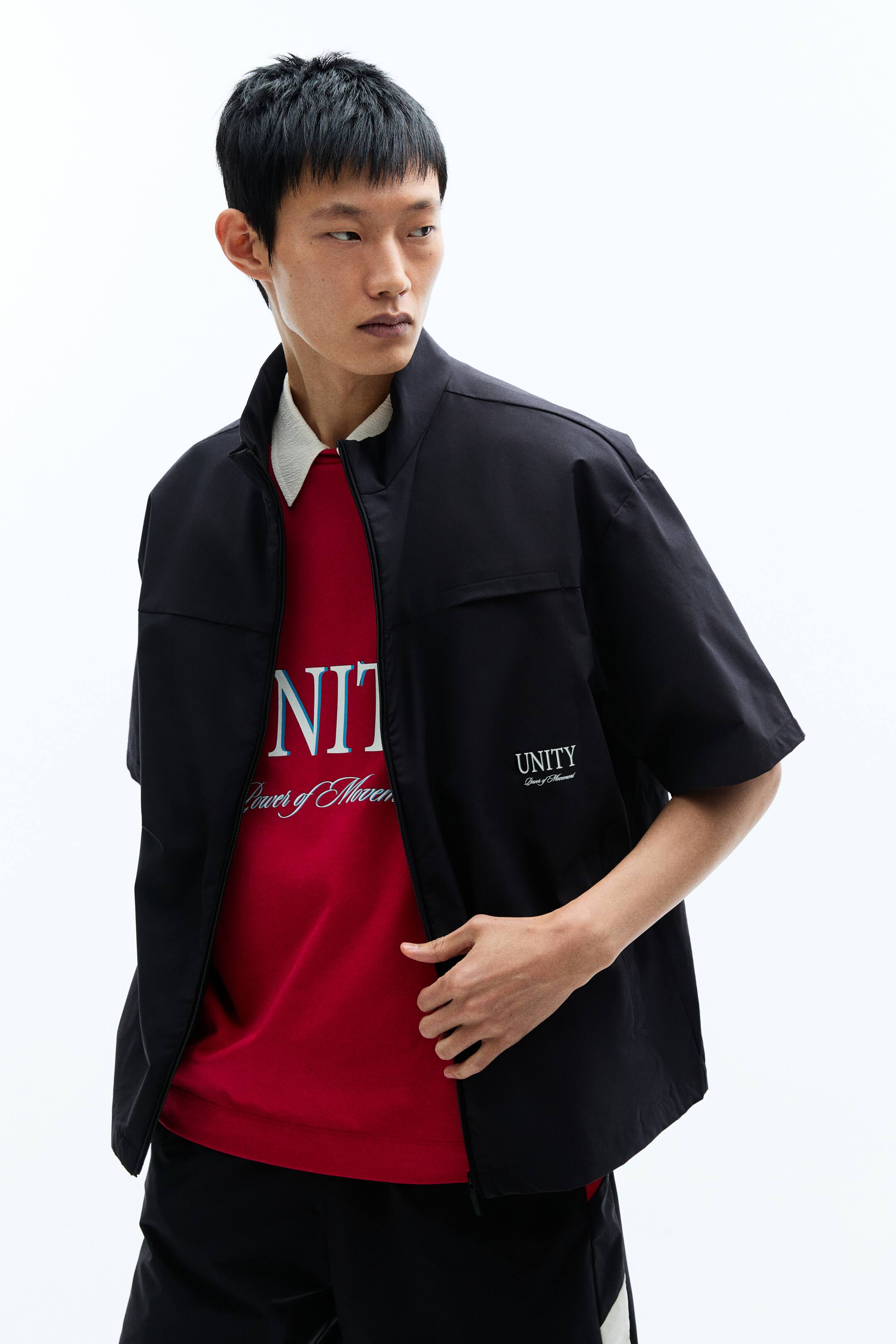 View larger image: A black short-sleeved jacket features a stand collar, a full-zip front and a relaxed fit, photographed facing slightly to the right. It shows two horizontal chest flap pockets and side pockets, with white embroidered text on the left chest, worn open over a red T-shirt.