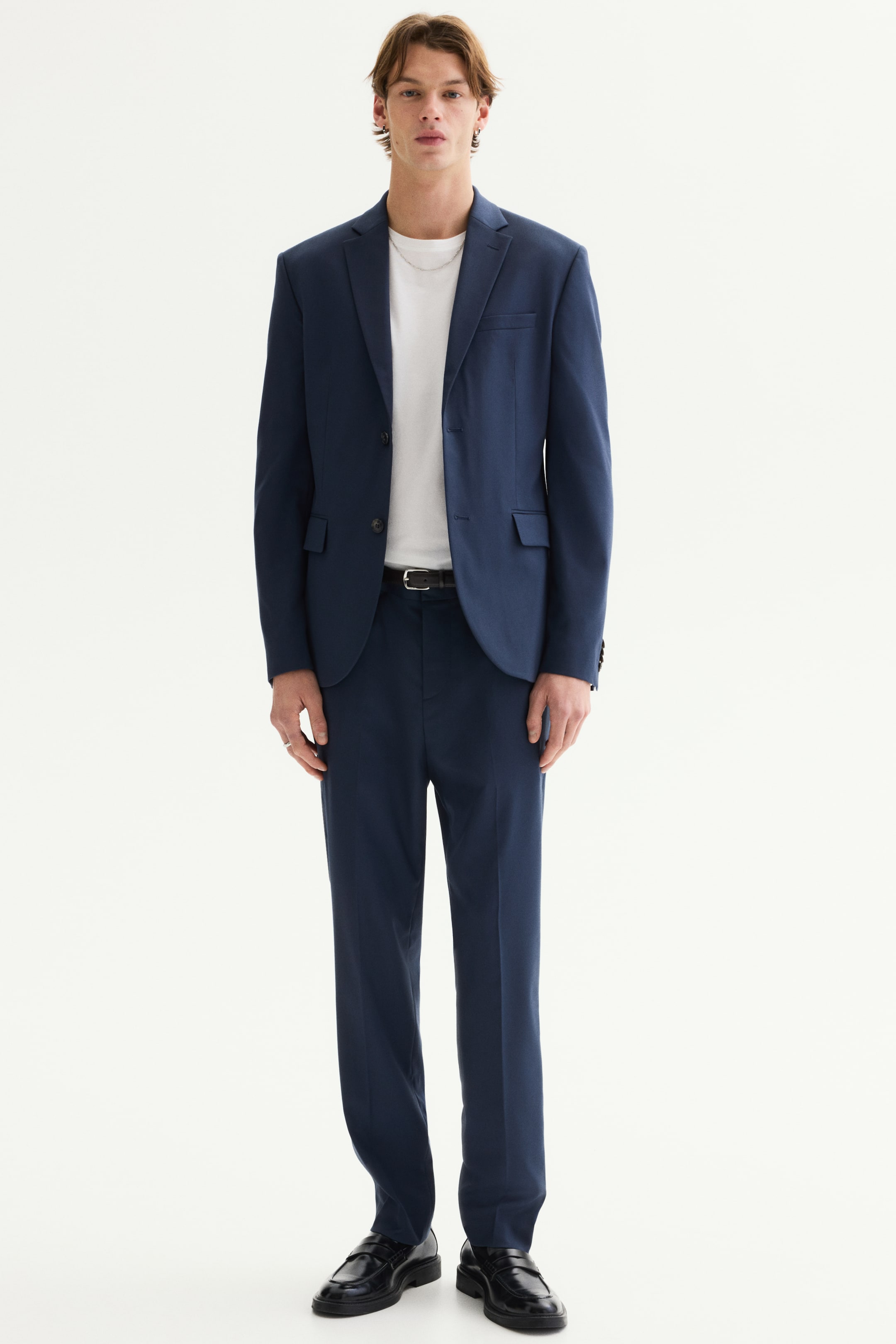 Преглед на по-голямо изображение: Тъмносин костюм е представен, обърнат директно напред, с двуредно сако с назъбени ревери, два джоба с капаци, джоб на гърдите и четири копчета на всеки маншет. Съответстващите панталони с права предна част са с прави крачоли, а черен колан е прокаран през халките за колан.