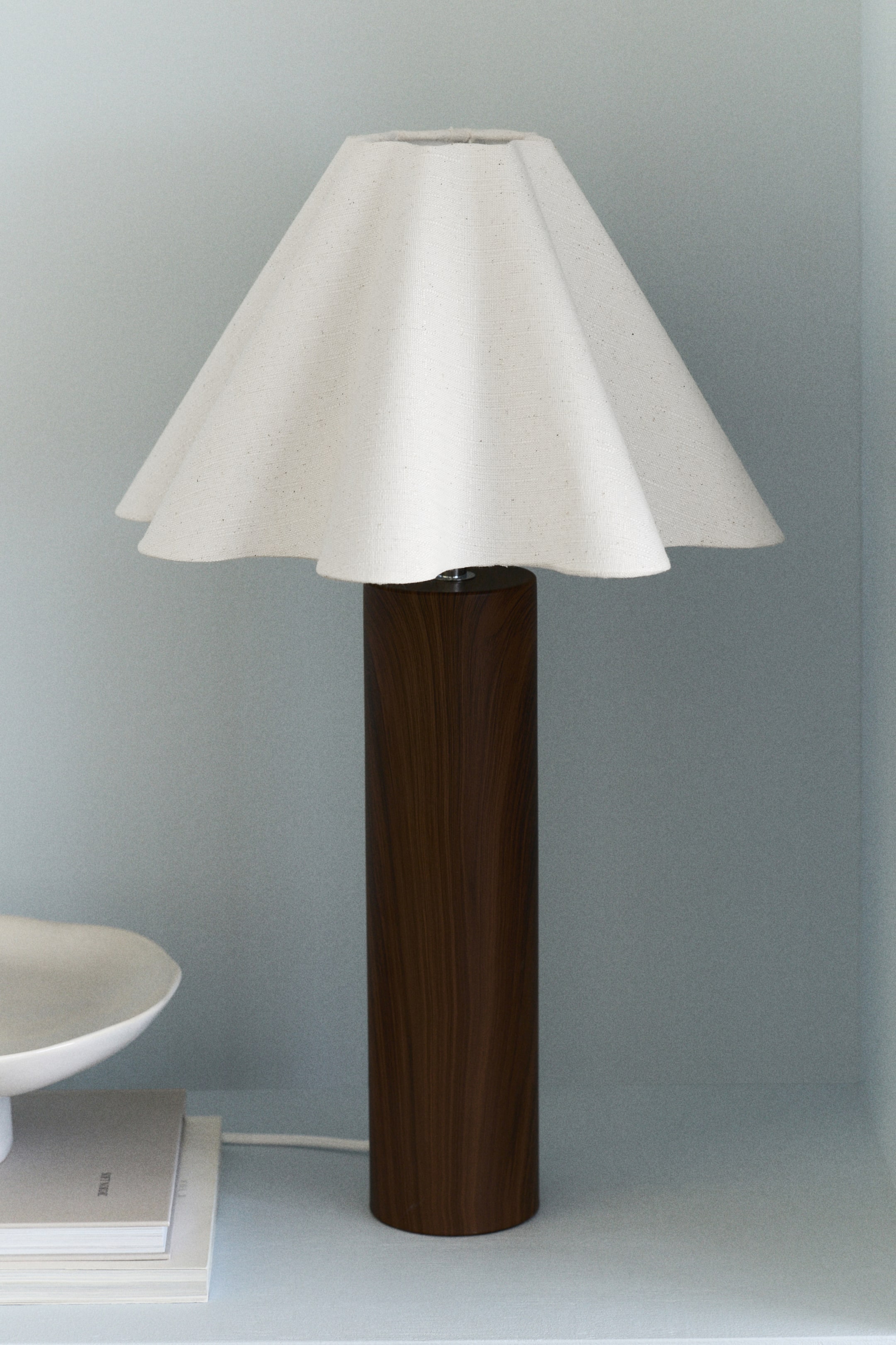 View larger image: A table lamp featuring a dark brown cylindrical base with a wood grain texture and a white power lead extending from its bottom. An off-white fabric shade flares outwards from the top of the base, showcasing a gently ruffled bottom edge.