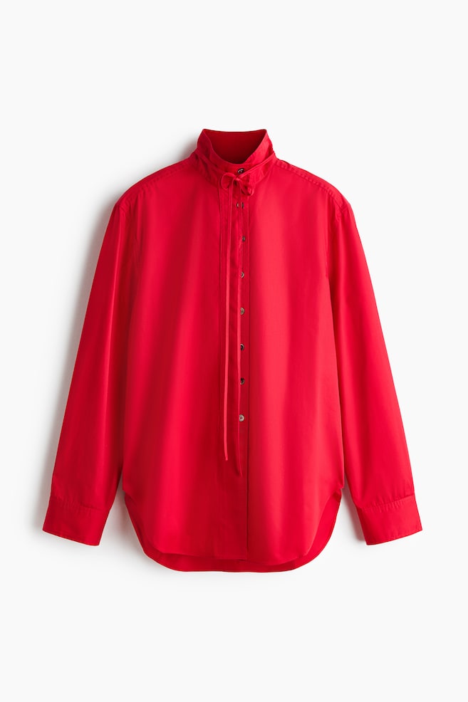 Una blusa roja se muestra en vista frontal, con un cuello alto adornado con un lazo decorativo anudado en el cuello, cuyos extremos caen sobre una tapeta de botones en el frente, mangas largas con puños de un solo botón y un dobladillo curvado. - 1