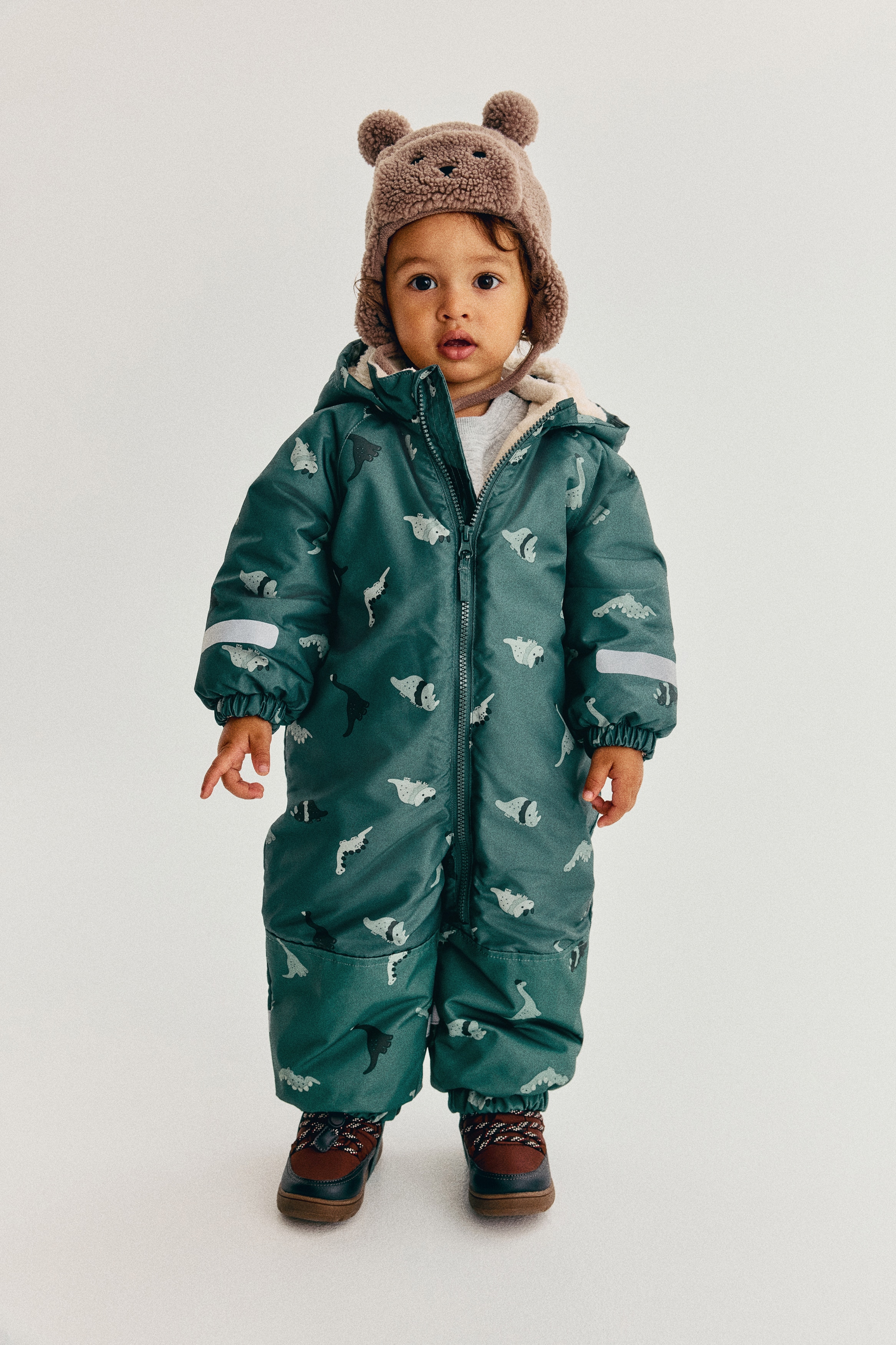 A padded all-in-one suit is showcasing its front, featuring a teal colour with an all-over dinosaur print. It is crafted with a padded hood featuring a light-coloured lining, a stand-up collar, and a full-length zipper running down the front. The suit includes elasticated cuffs, elasticated hems with straps extending under the feet, reinforced knee sections, and reflective strips on the arms.