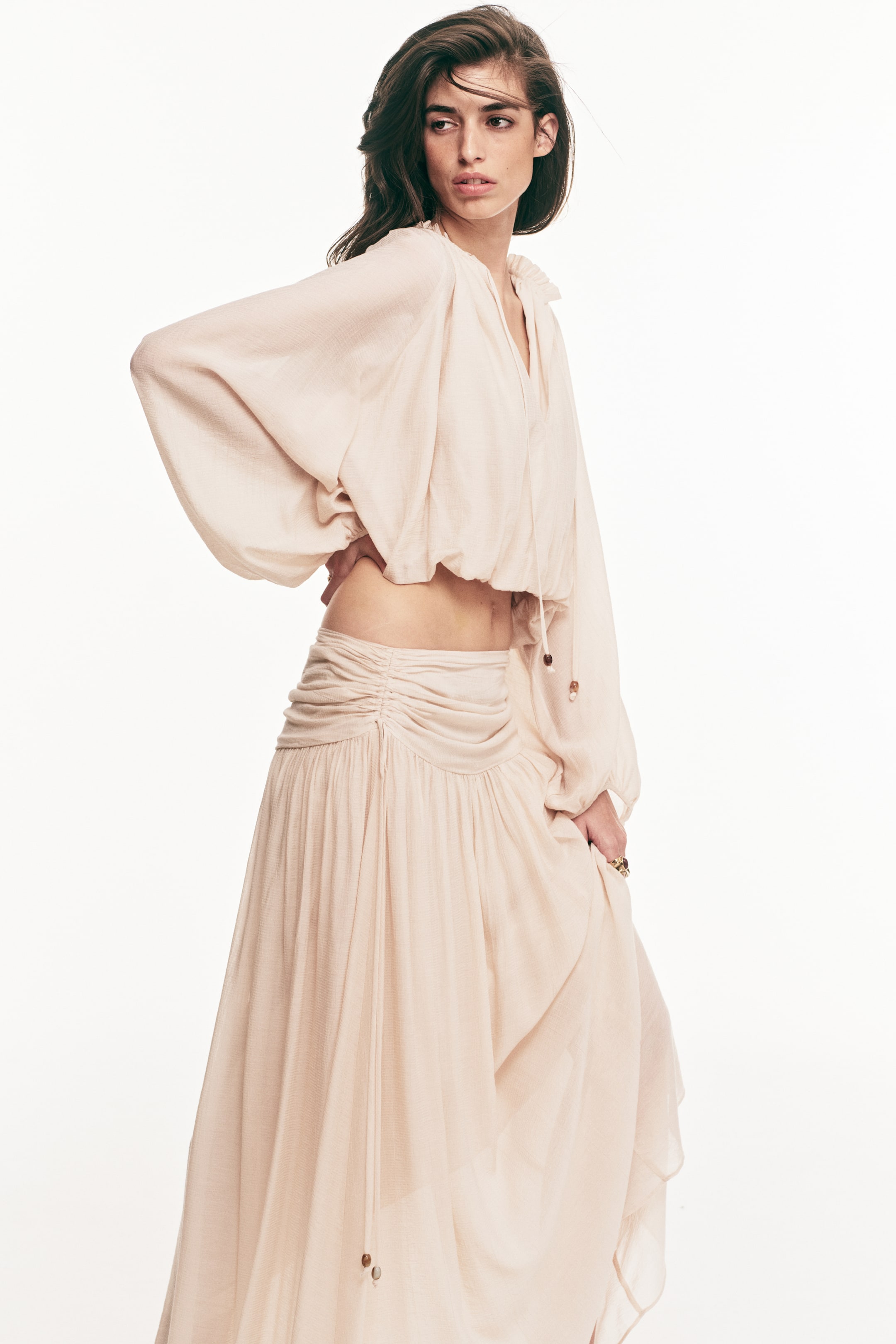 View larger image: A light beige two-piece outfit is showcased from a three-quarter left angle, comprising a cropped blouse and a flowing maxi skirt. The blouse boasts a ruffled V-neckline, voluminous long sleeves, and a gathered elasticated hem with drawstrings and beads, while the skirt features ruched detailing at the waist and a flowing hem, also adorned with a drawstring and beads.