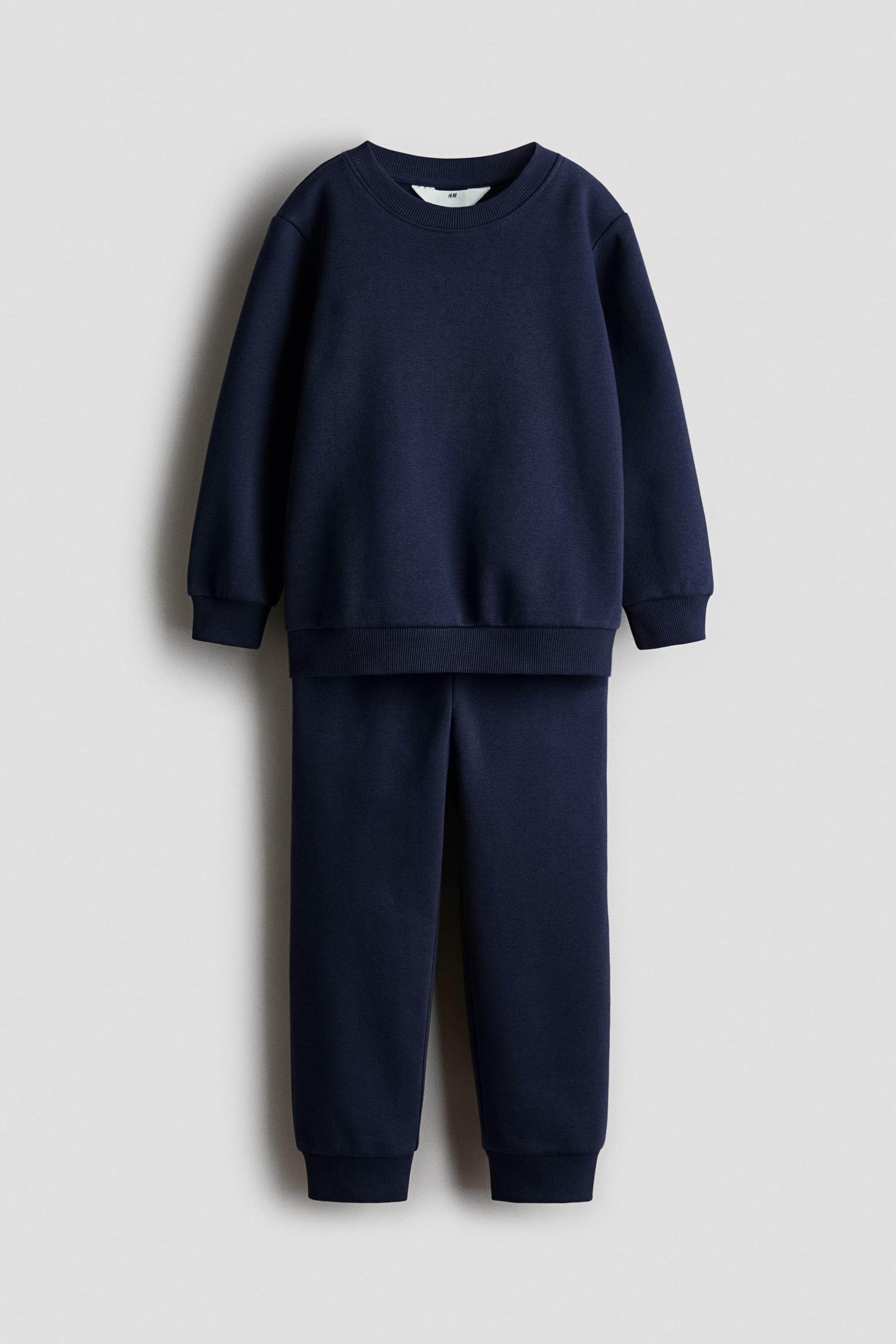 Größeres Bild ansehen: Ein marineblaues Trainingsanzug-Set, von vorne betrachtet, bestehend aus einem Rundhals-Sweatshirt und passenden Jogginghosen. Das Sweatshirt verfügt über einen gerippten Rundhalsausschnitt, gerippte Bündchen und einen gerippten Saum, und die Jogginghosen haben ebenfalls gerippte Bündchen an den Knöcheln.