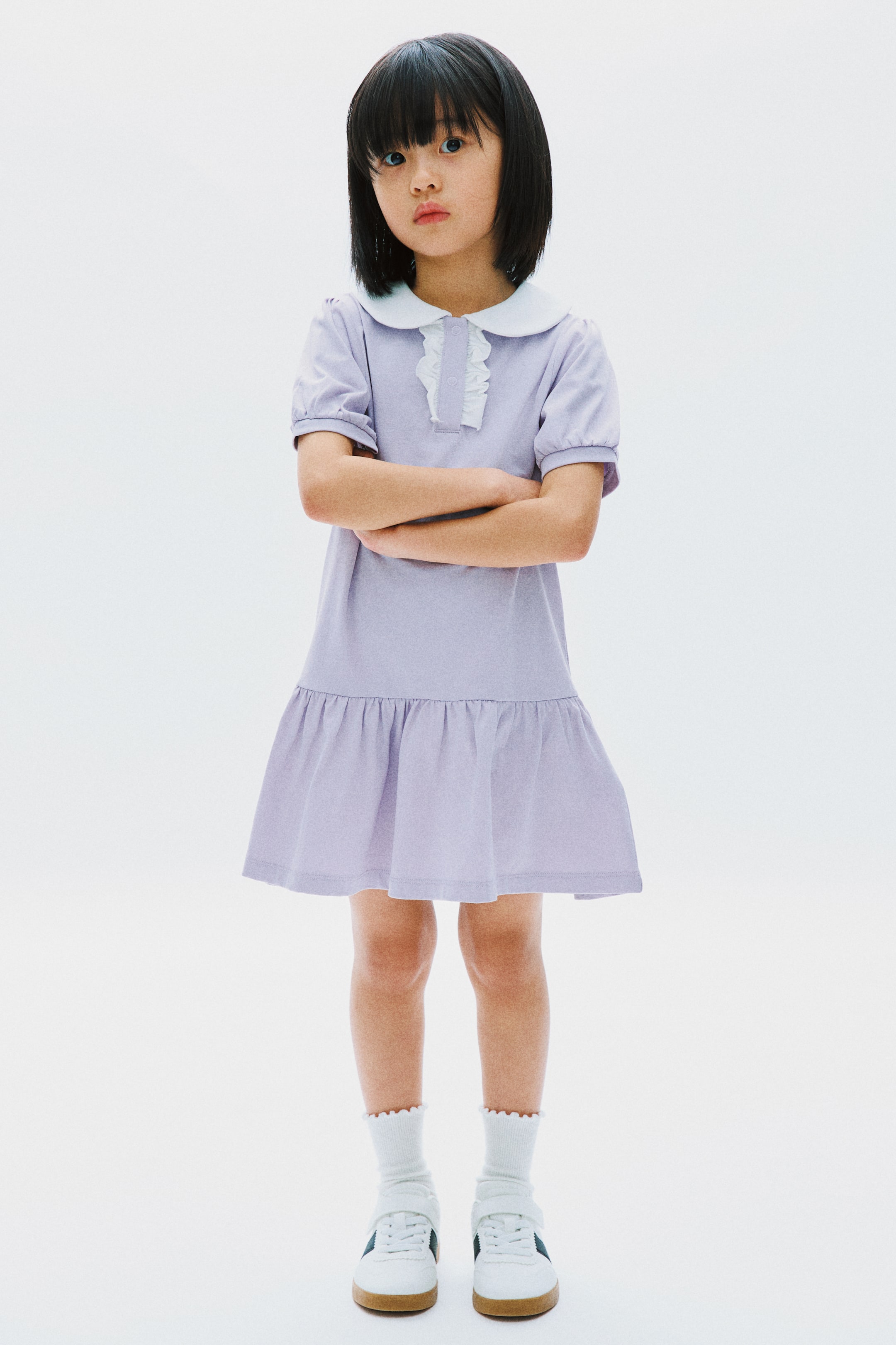 Xem ảnh lớn hơn: Một chiếc váy màu oải hương phía trước nổi bật với cổ áo Peter Pan tròn màu trắng và diềm xếp nếp trắng dọc theo giữa phía trước. Nó được thiết kế với tay áo ngắn phồng, bó lại bằng cổ tay đàn hồi, và váy xếp tầng tụ lại phía trên gấu.