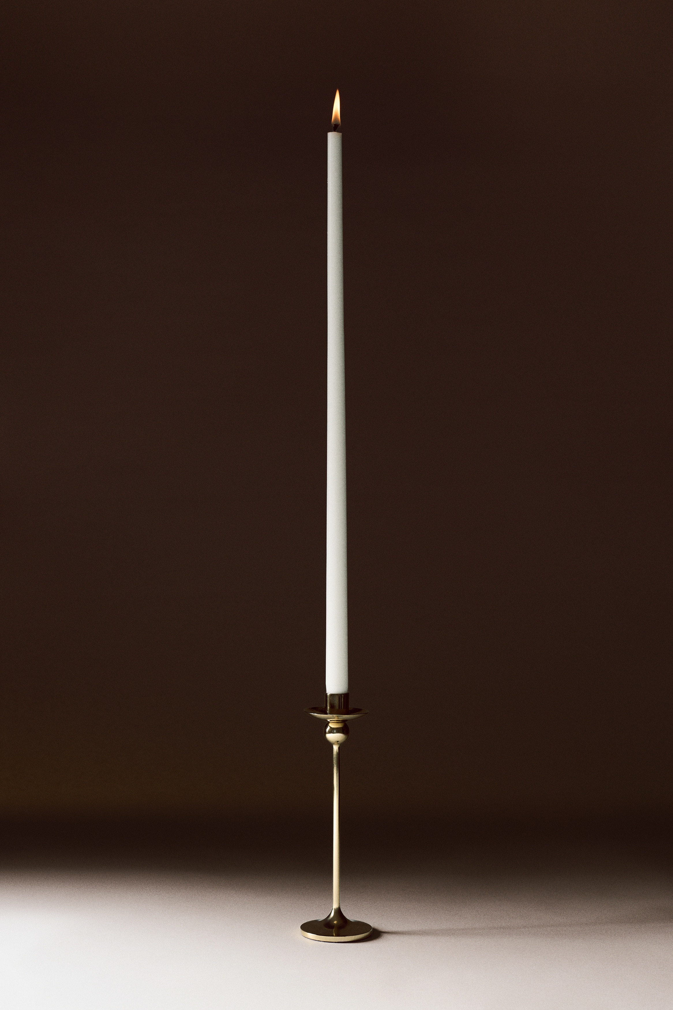 A polished stainless steel candlestick with a gold finish stands upright, featuring a sleek, slender stem rising from a flared circular base. The design includes a spherical detail positioned directly beneath a shallow drip tray and a cylindrical candle holder at the top.